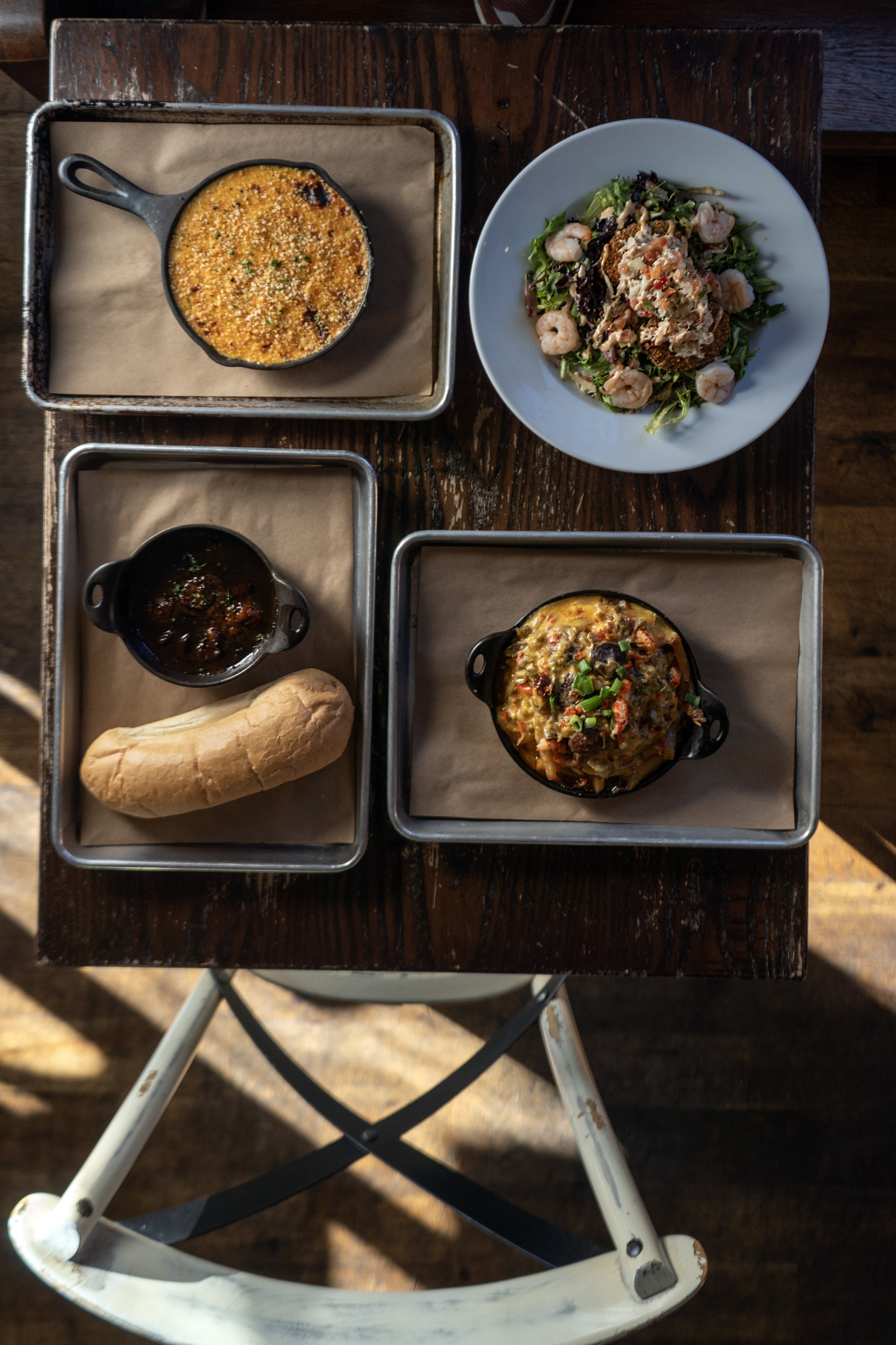 A top-down view of a rustic wooden table with a meal including a white plate with salad and shrimp, two small black cast-iron dishes with baked dishes, a small black cast-iron bowl with sauce, a bread roll, and an empty baking tray.