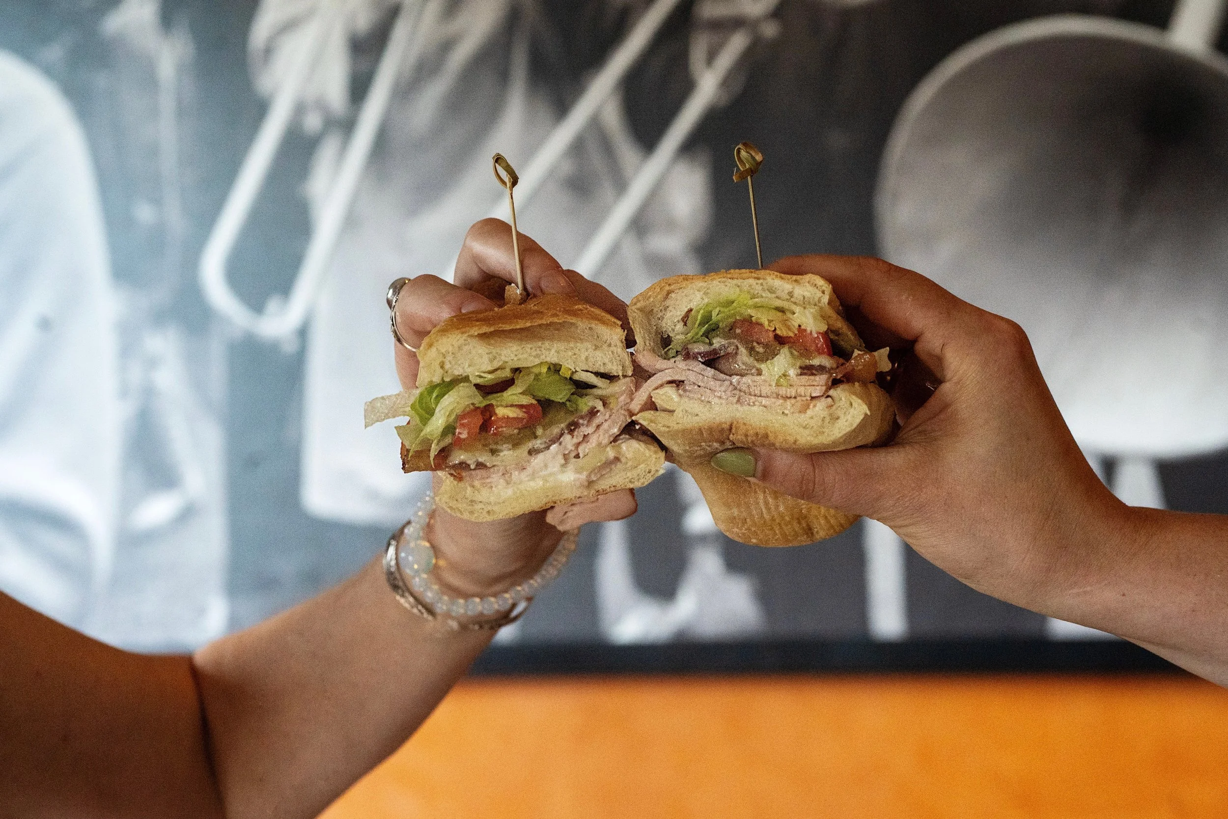 A person holds a sliced poboy sandwich with lettuce, tomato, sliced turkey, and other vegetables in a bun, with a background of new orleans jazz musicians