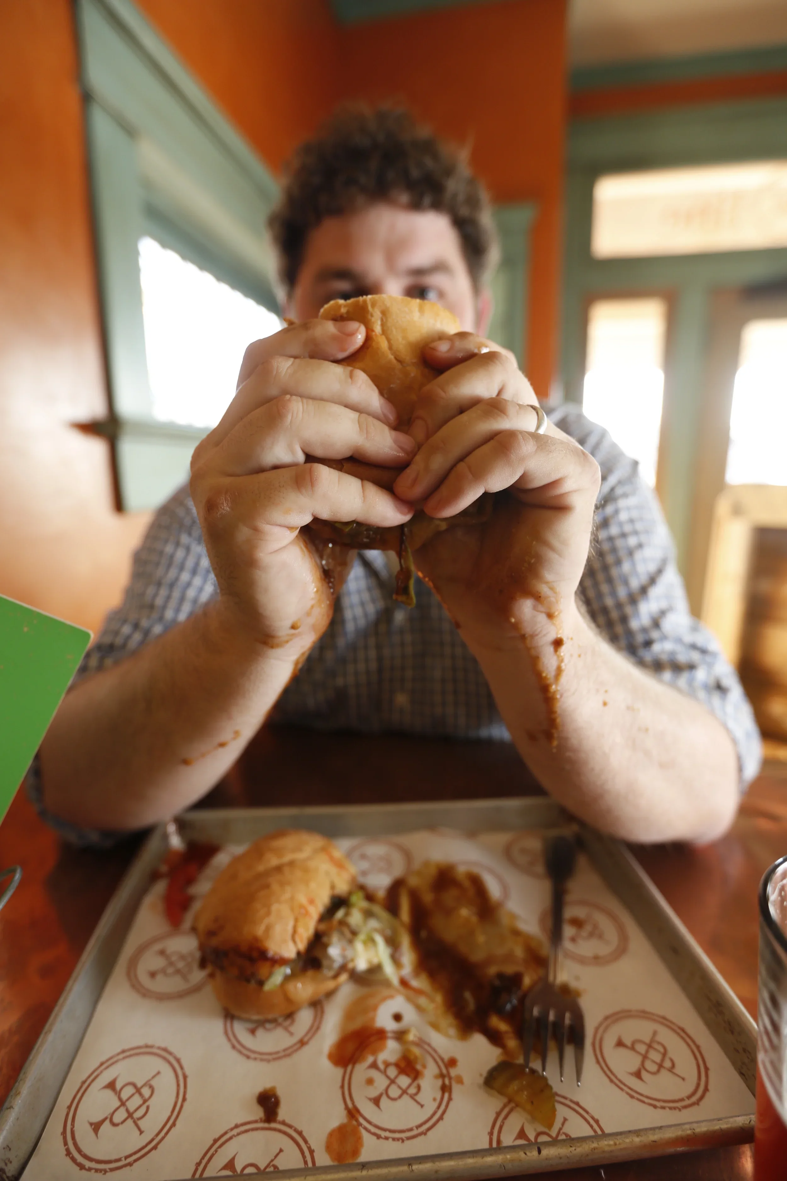 Chef Kelly English holding a roast beef poboy with both hands, sitting at a table with a partially eaten oast beef poboy  and tray, in a restaurant with wooden walls and windows.