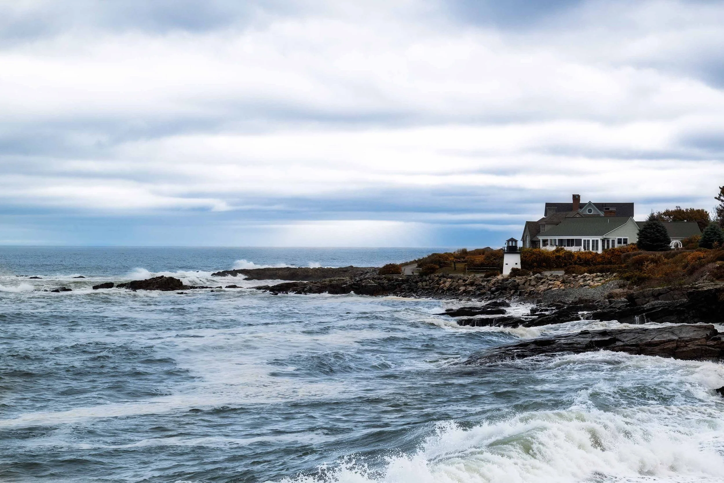 View of Lobster Point Lighthouse along Marginal Way in Ogunquit, Maine, with waves crashing against rocky shoreline and a coastal cottage under dramatic storm clouds, captured as fine art wall art.