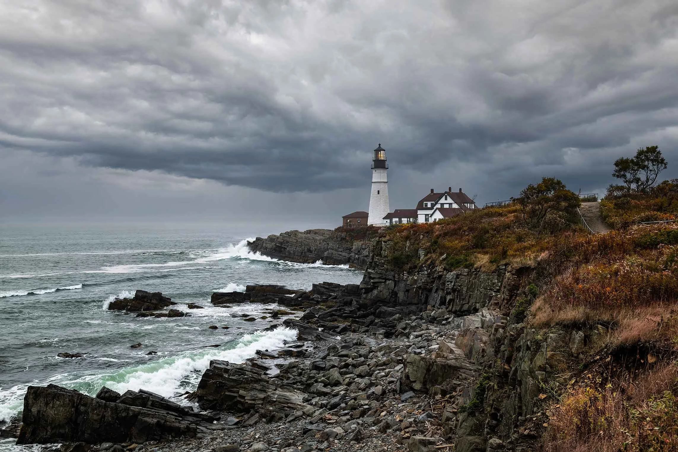 Portland Head Light in Cape Elizabeth, Maine with waves crashing against the rocky coast as a storm approaches, captured as dramatic coastal fine art photography for buyers seeking Maine lighthouse wall art.