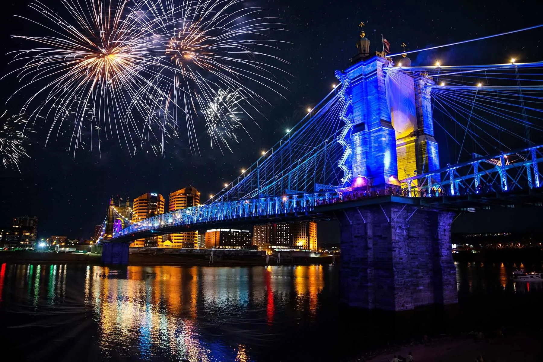 John A. Roebling Suspension Bridge in Cincinnati dressed in blue light for BLINK, fireworks exploding over the bridge and the Cincinnati skyline at night.