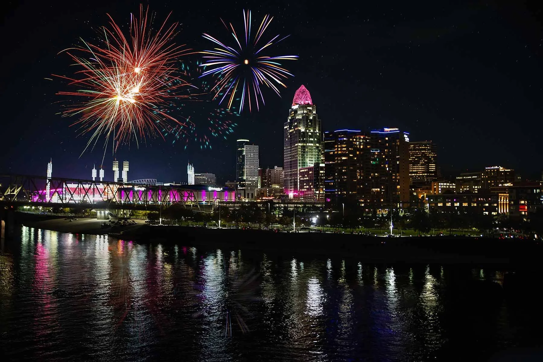 Night photo of the Cincinnati skyline during Light Up Cincinnati, with downtown towers and riverfront buildings illuminated against the dark sky. Fireworks decorating the skyline.