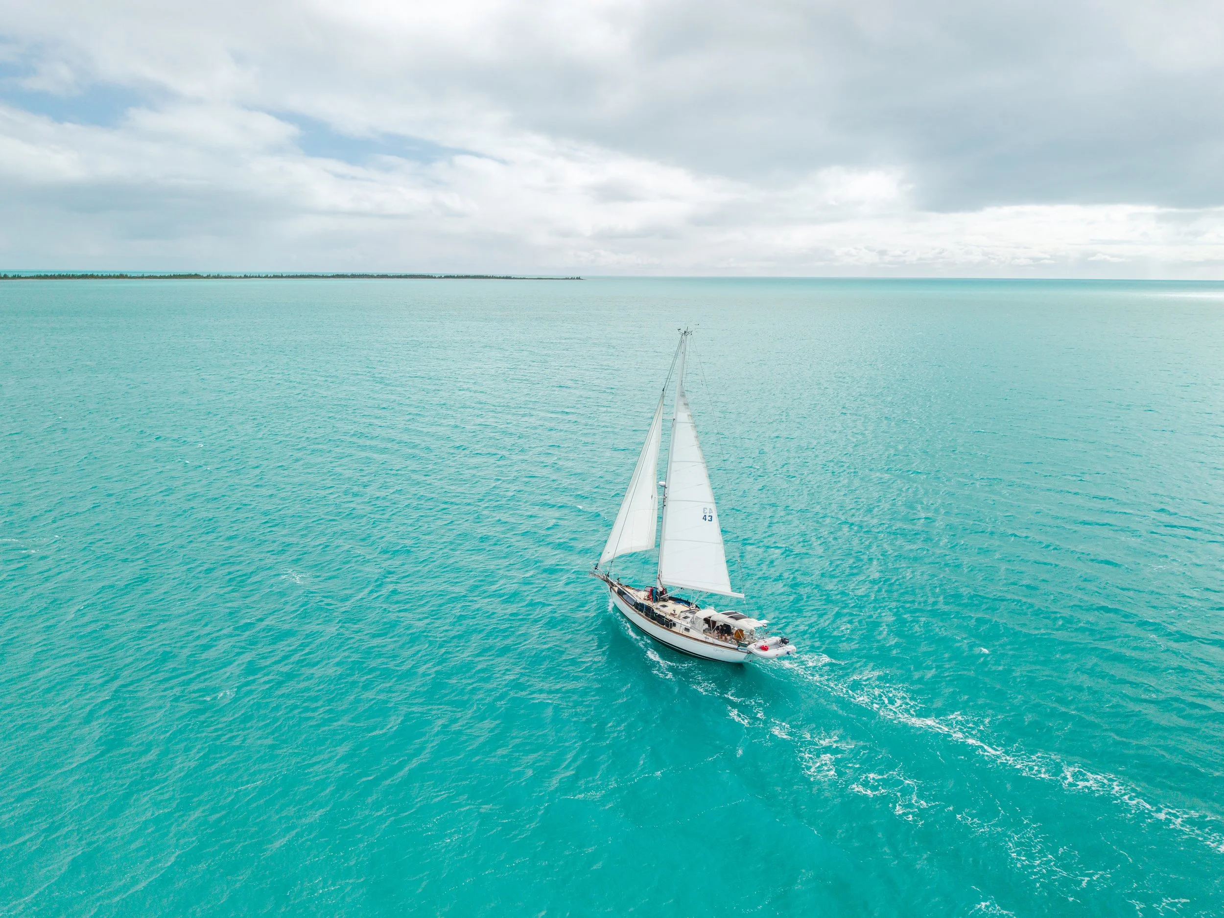 A sailboat with white sails on calm, turquoise waters under a partly cloudy sky, with land visible in the background.
