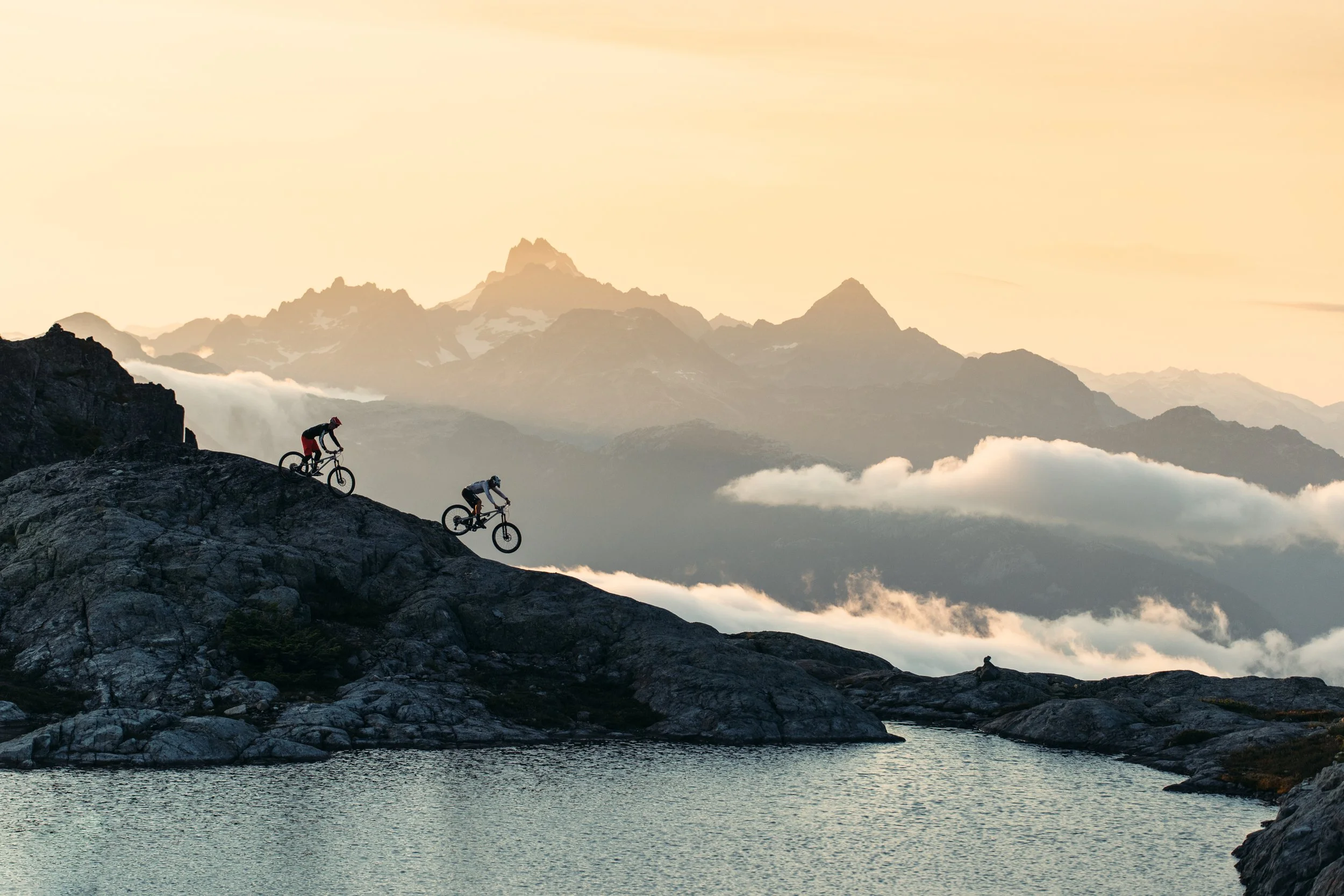 Two mountain bikers riding on rocky terrain near a body of water, with a backdrop of high mountain peaks and a soft, cloudy sky at sunset.