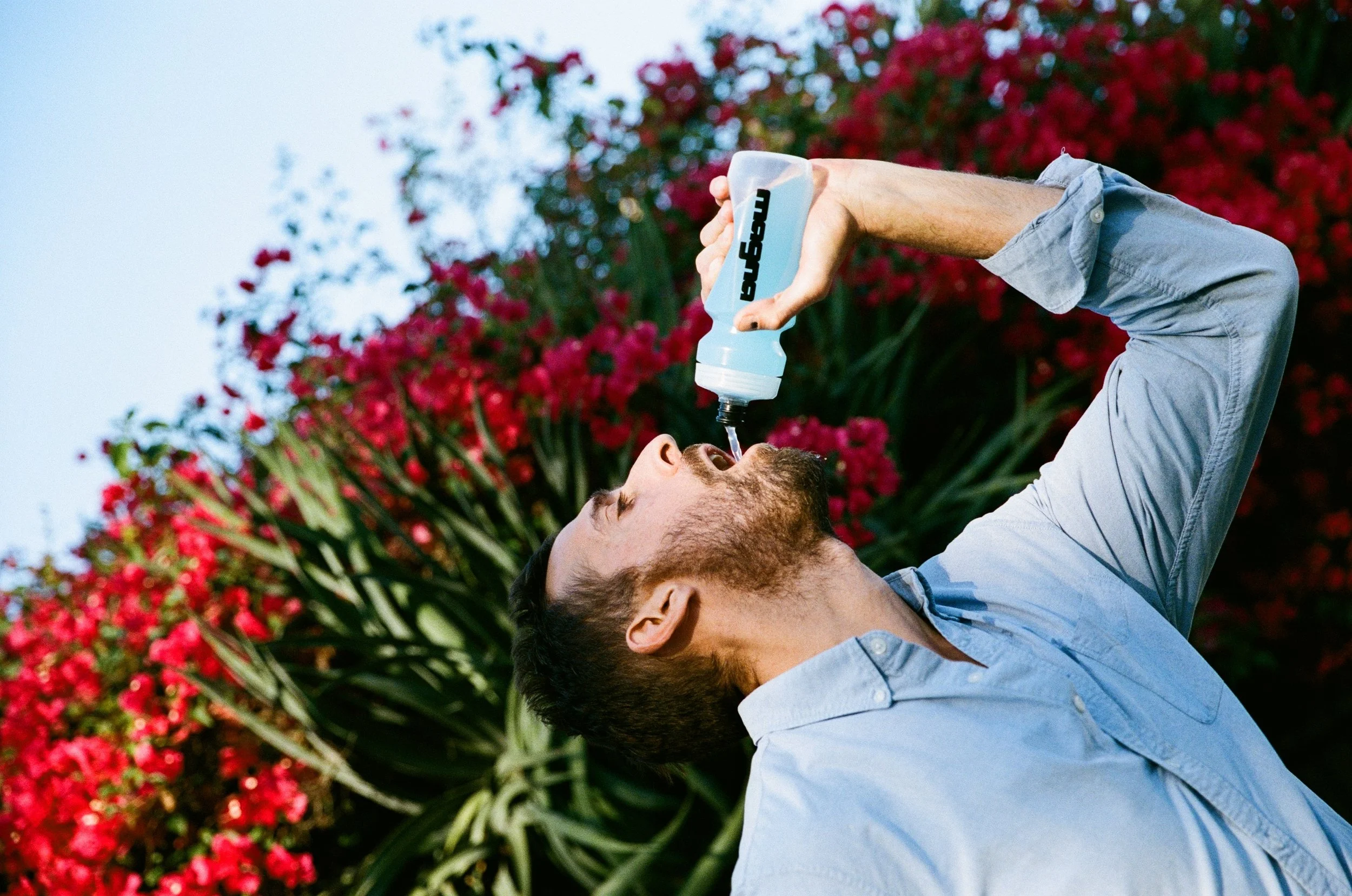 A man lying down outdoors, drinking water from a bottle labeled 'BEACH' with a background of vibrant pink and red flowers and green foliage.
