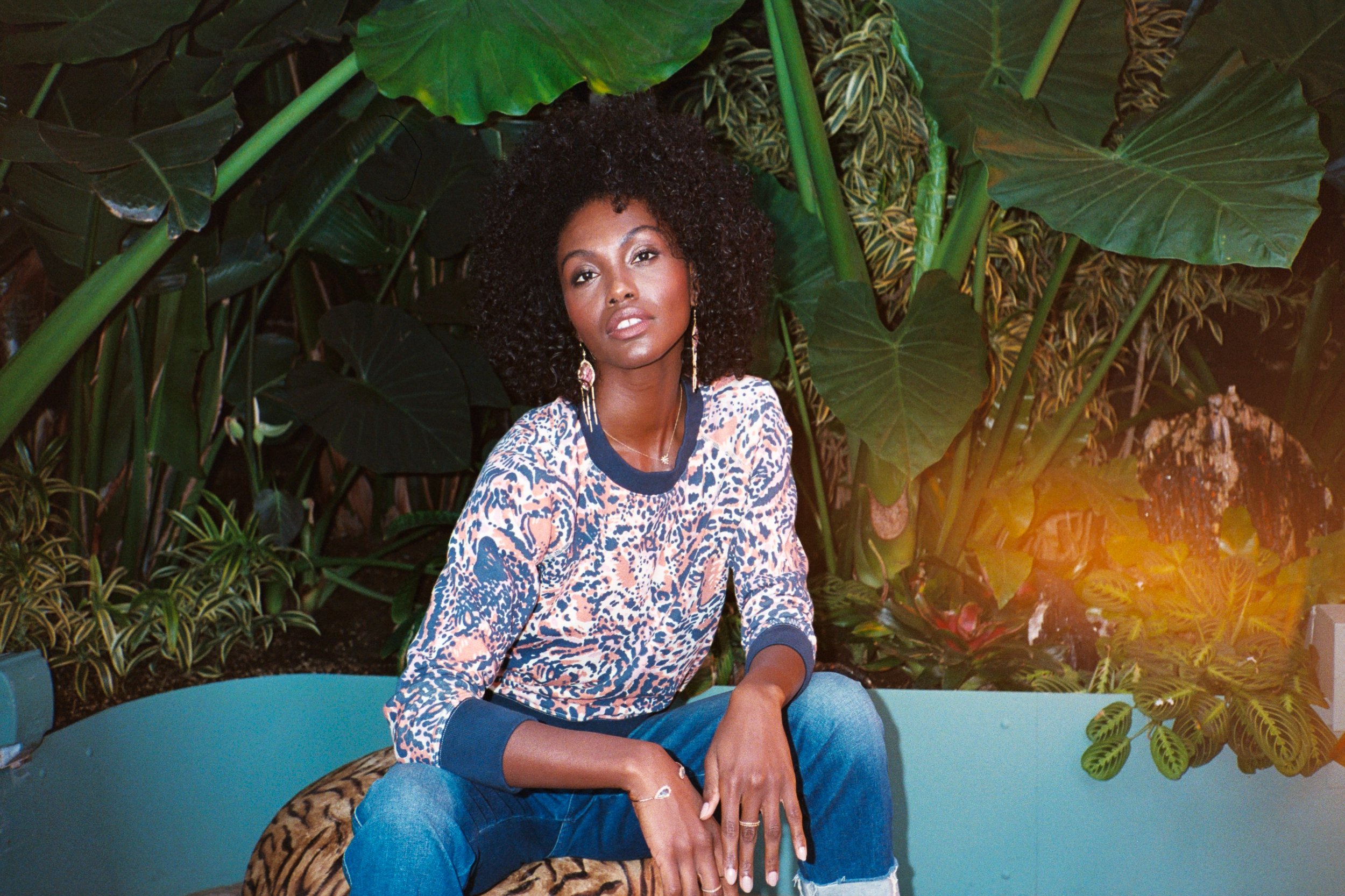 A woman with curly hair sitting outdoors in front of lush green plants and large leaves, wearing a patterned top and ripped jeans.