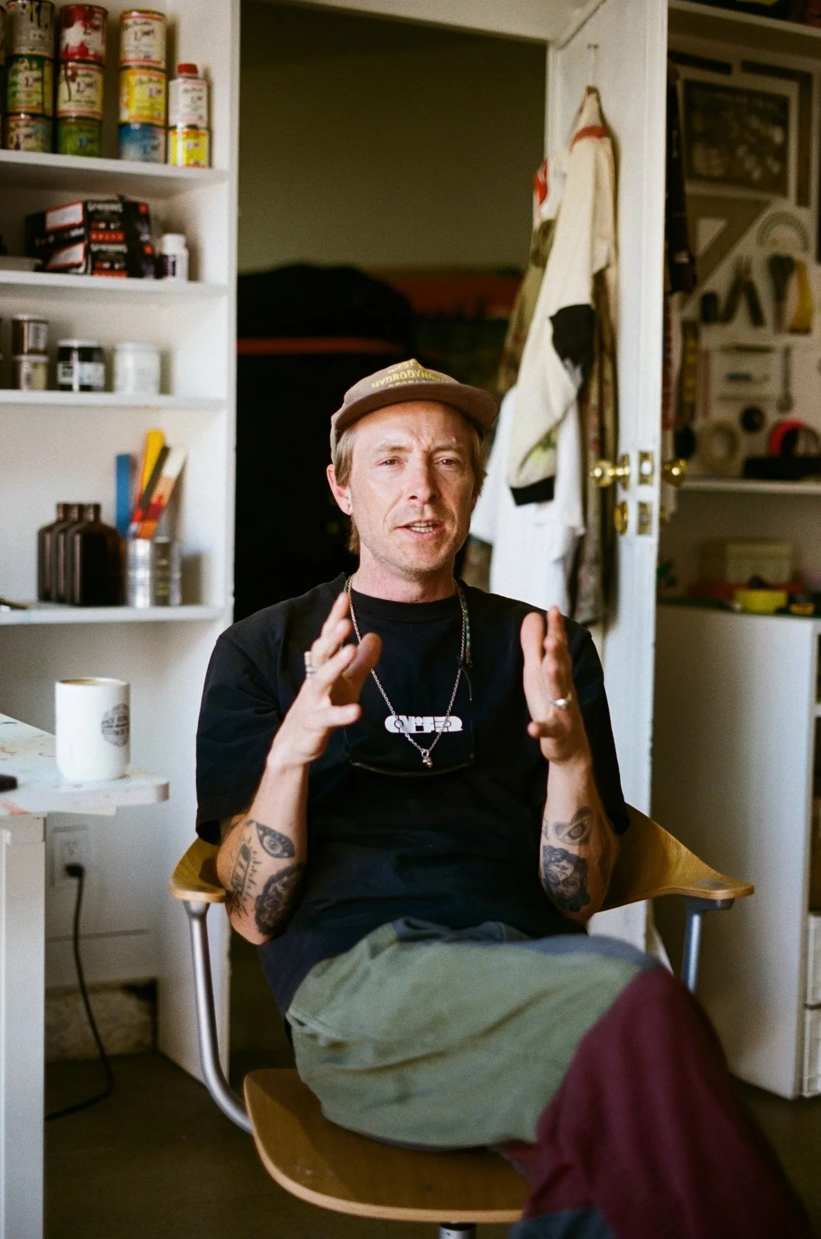 A man with tattoos on his arms, wearing a brown cap and black T-shirt, sitting in a wooden chair, gesturing with his hands, inside a room with shelves of canned food and supplies in the background.