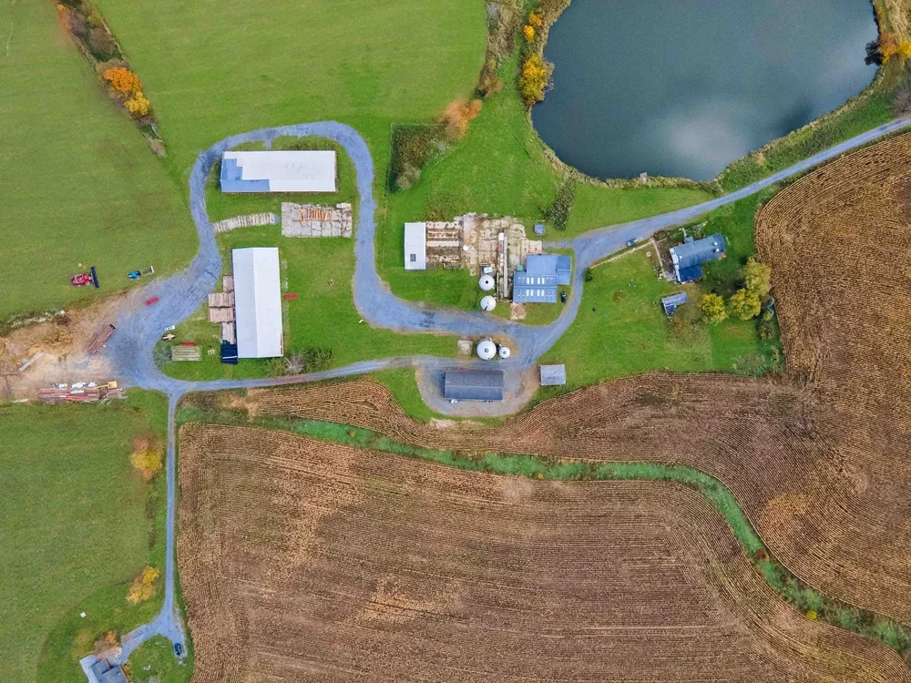 Aerial view of a farm with multiple buildings, a pond, and fields. Gravel paths connect the structures and fields.