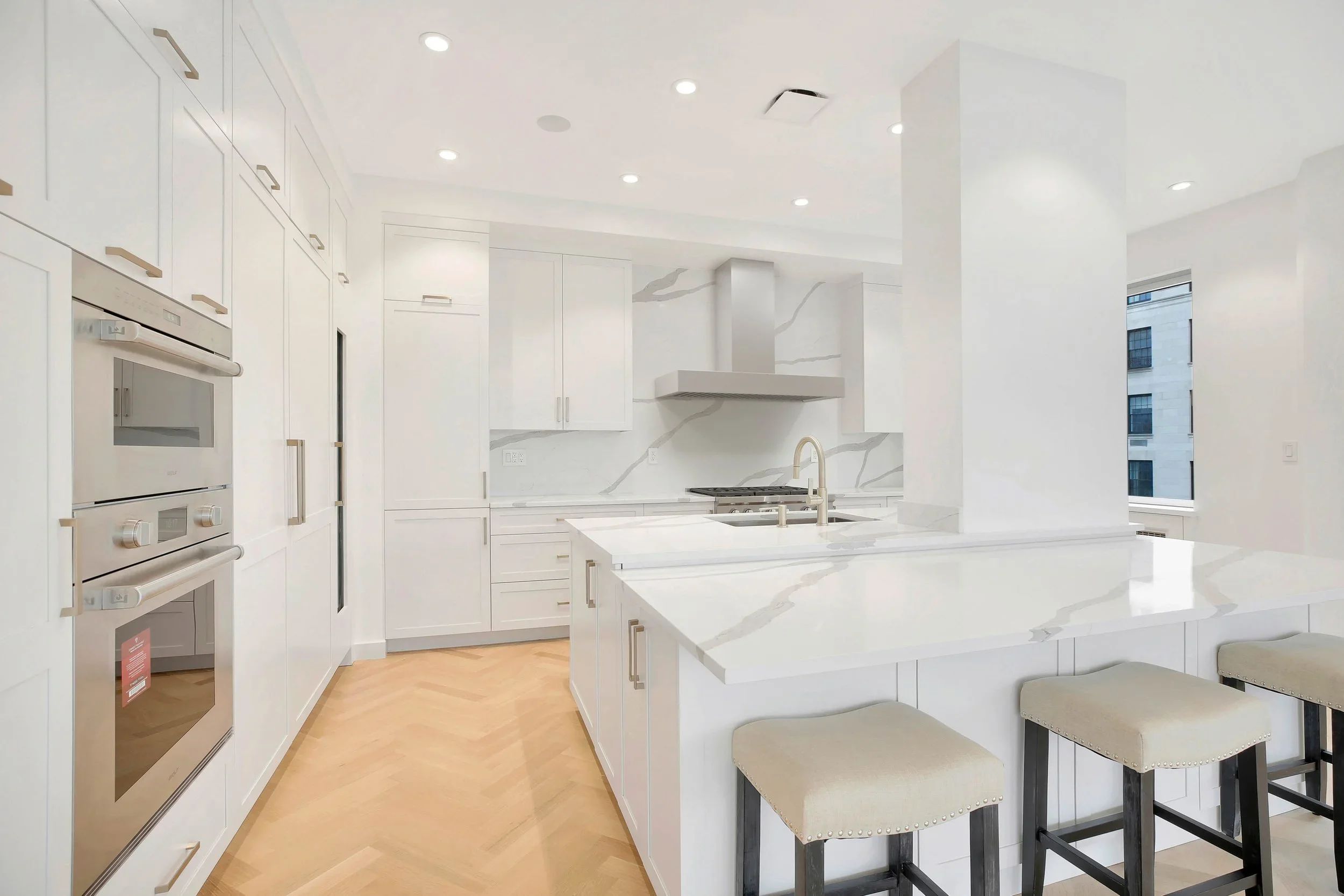 Bright white modern kitchen with marble countertops, built-in oven and microwave, large island with seating, and large windows showing city buildings outside.