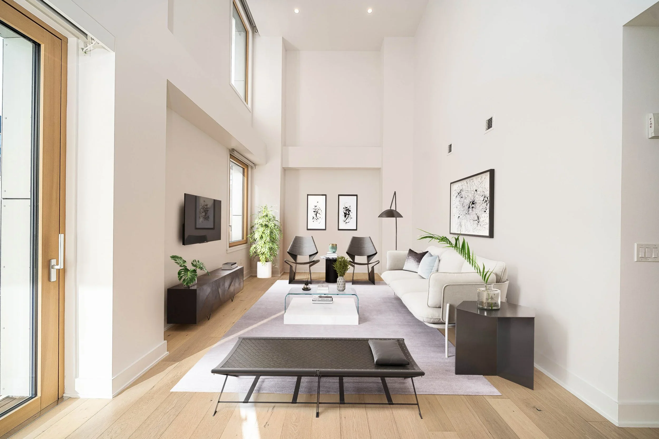 Modern living room with high ceilings, hardwood floors, white walls, large windows, a flat-screen TV, framed artwork, a white sofa, black chairs, a coffee table, a floor lamp, plants, and a patio door.