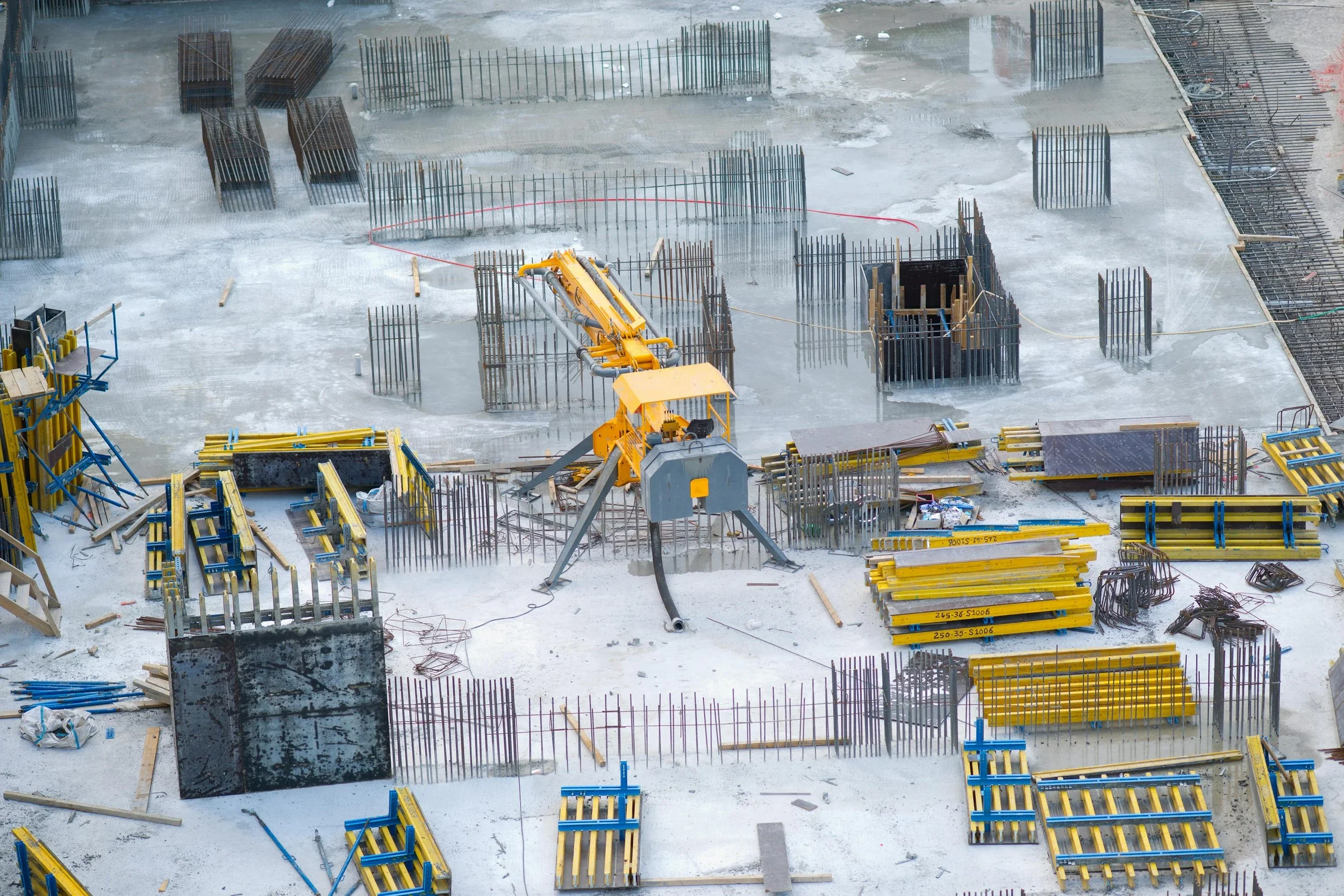 Construction site with numerous steel reinforcement bars, wooden forms, and yellow construction equipment on a wet concrete surface.