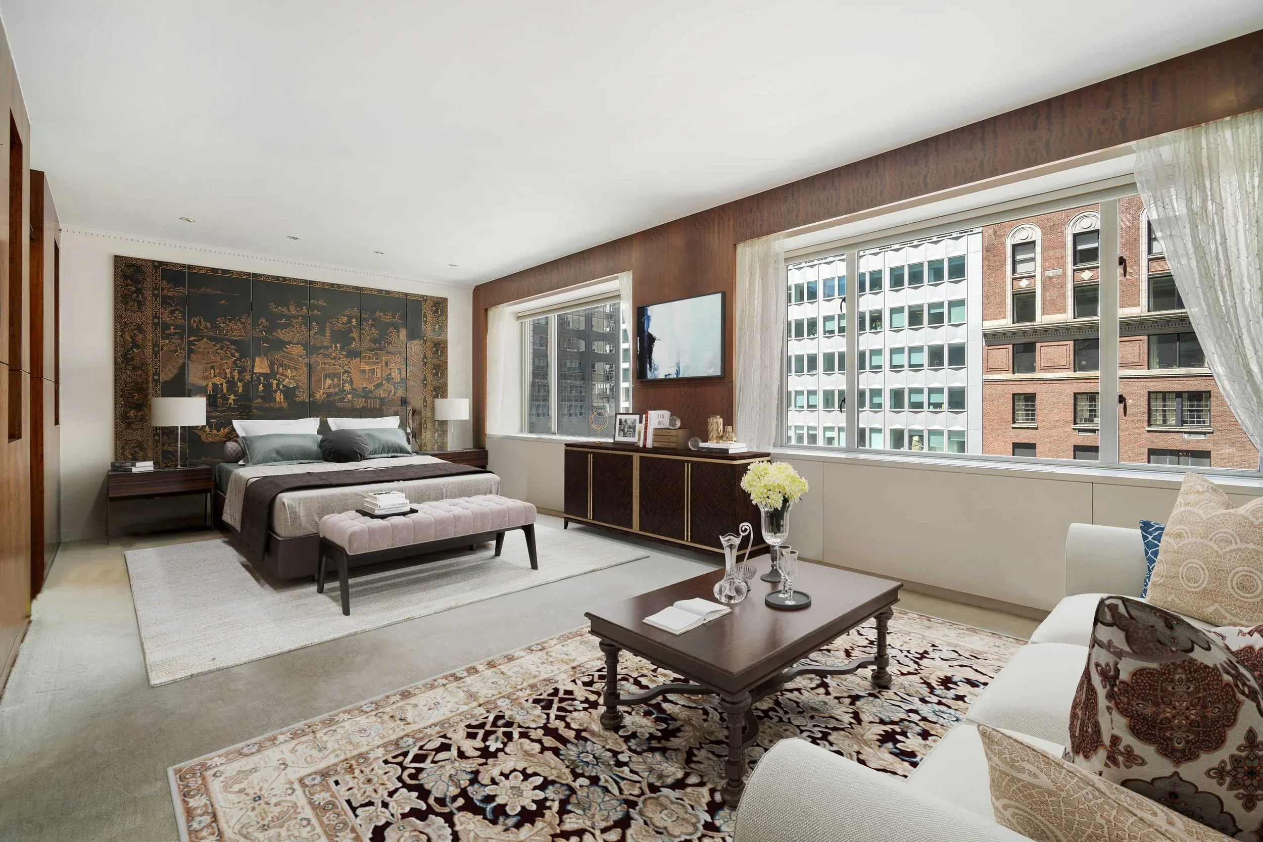 A spacious living room with large windows showing city buildings outside. The room features a bed with pillows, two side tables with lamps, a sofa, a coffee table with decorative glassware, and a Persian-style rug. There's a dark decorative screen wi