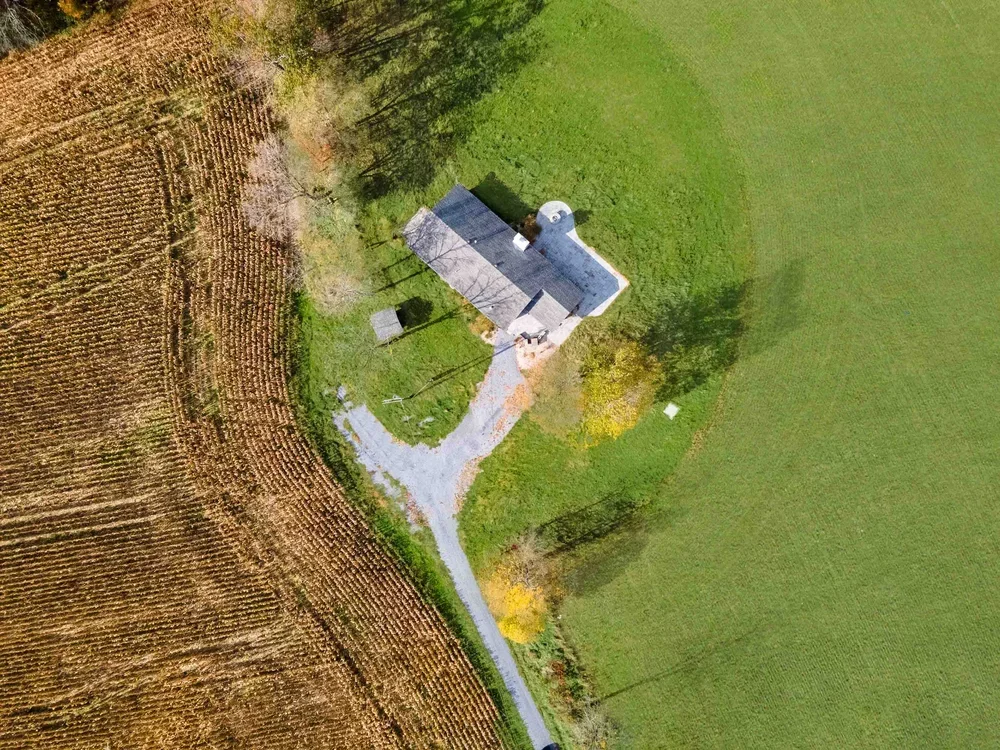 An aerial view of a house surrounded by green grass on one side and harvested farmland on the other, with a gravel driveway leading to the house.
