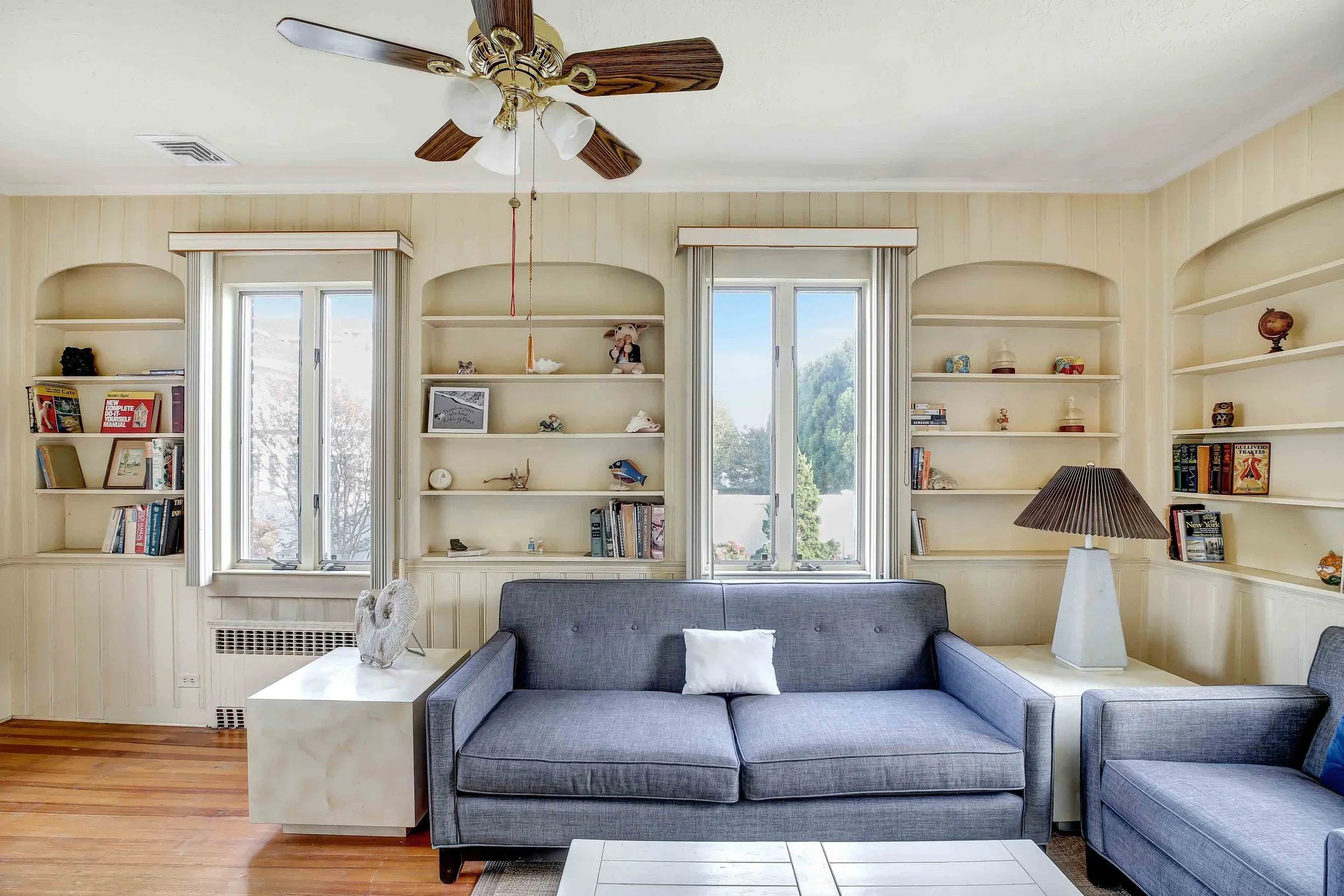 Living room with a ceiling fan, large windows, built-in bookshelves, a gray sofa, a white coffee table, and a table lamp.