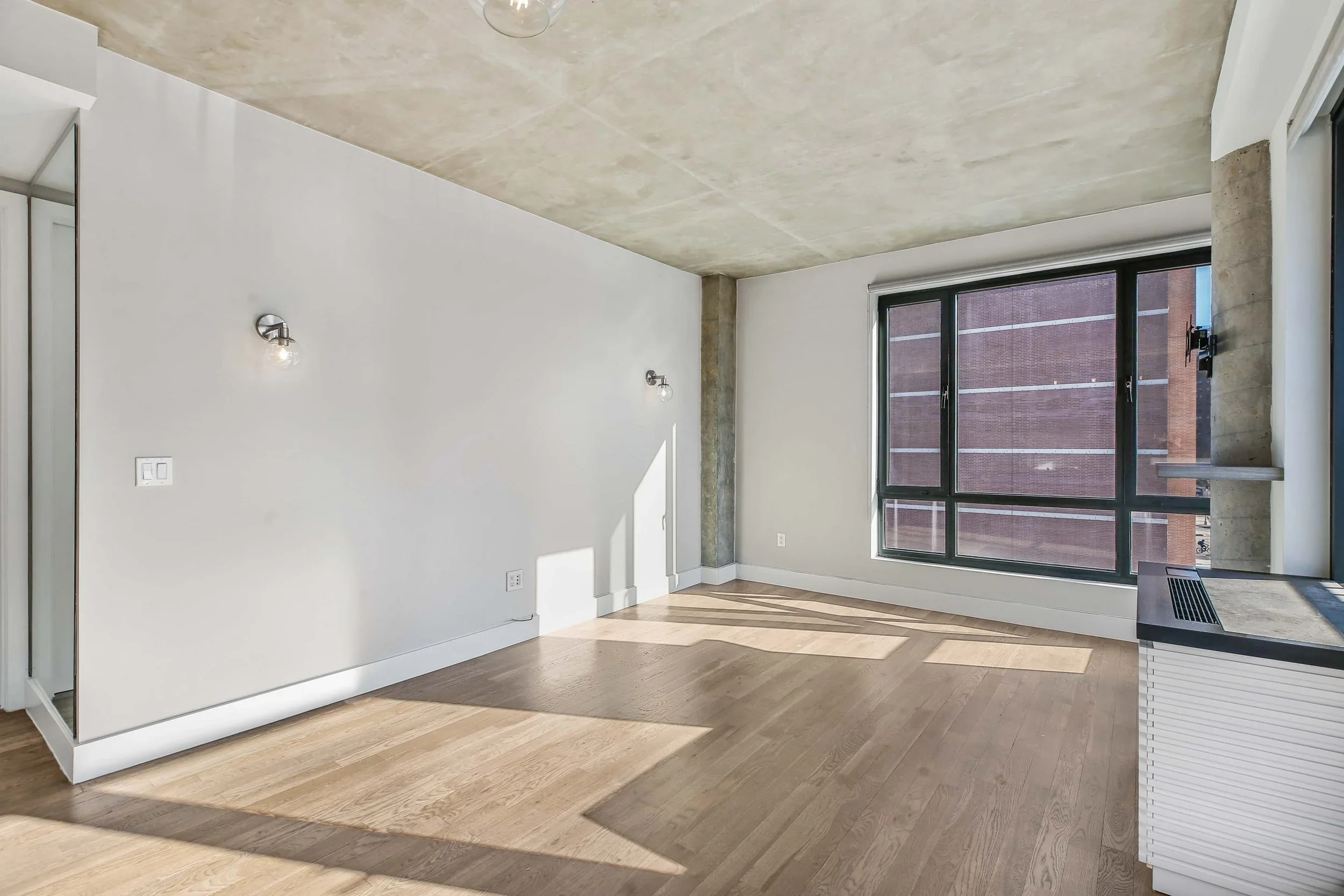 Empty room with wooden flooring, white walls, large window, and concrete ceiling.