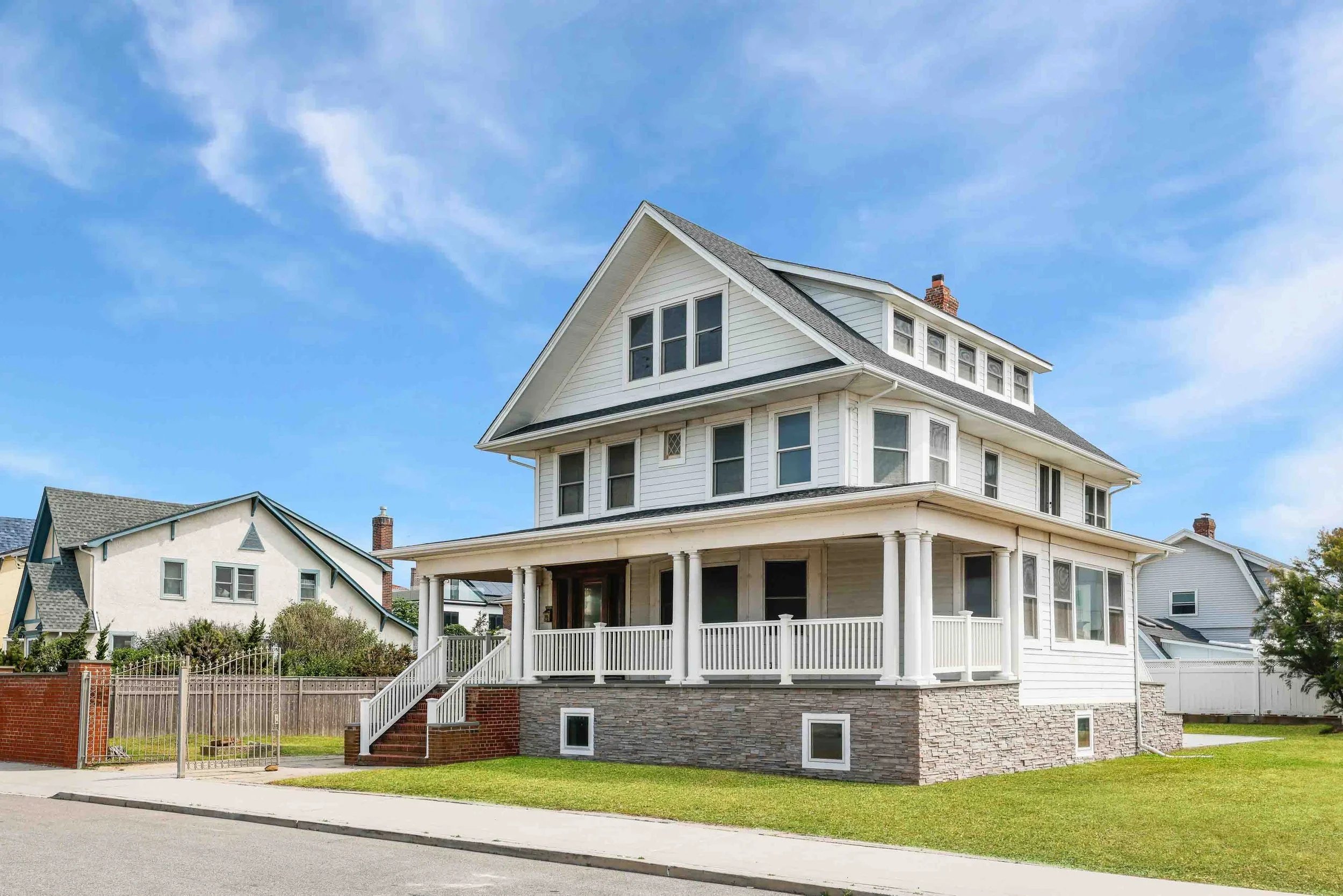 A large white three-story house with a front porch, multiple windows, and a fenced yard, under a partly cloudy blue sky.