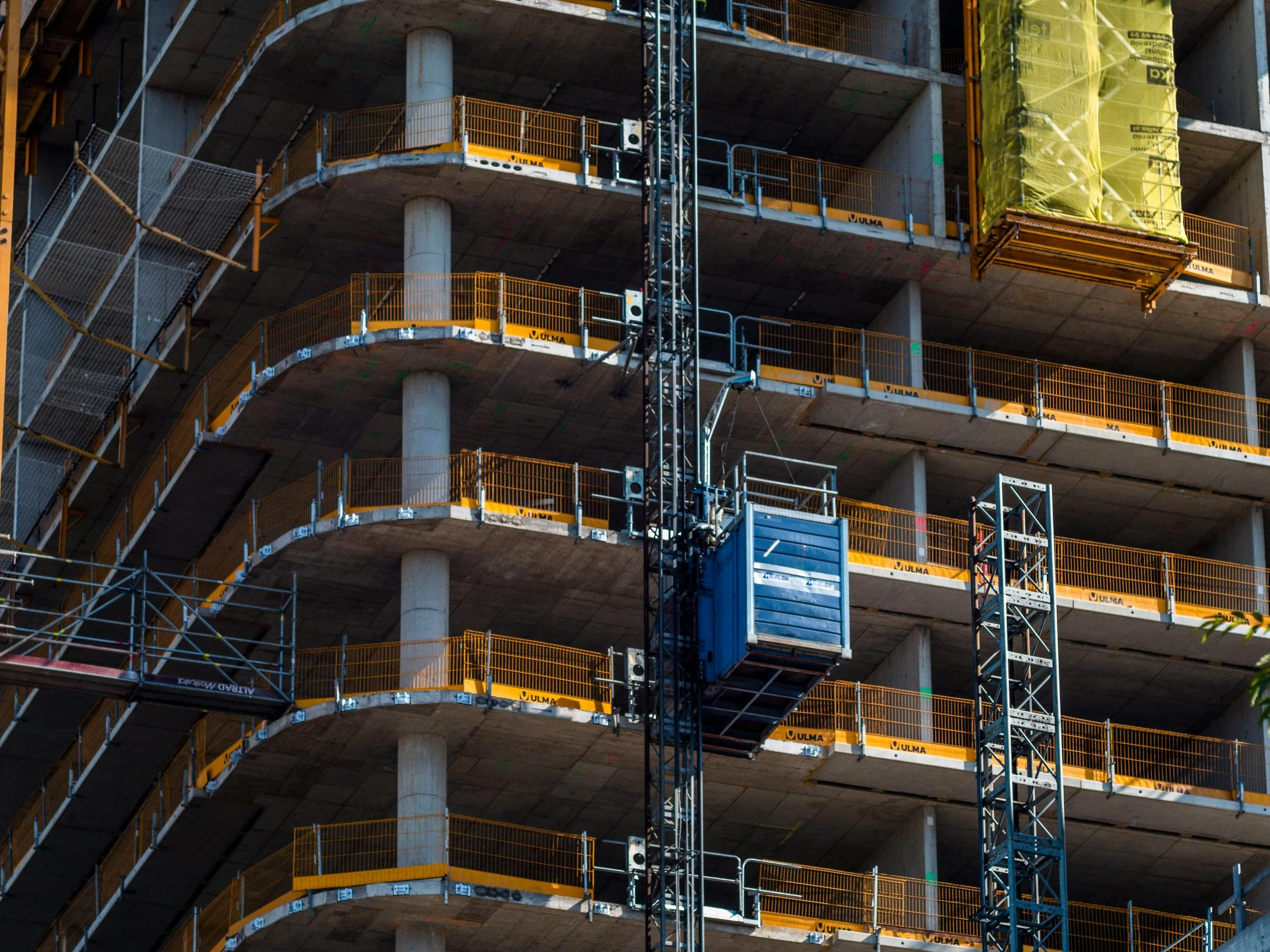 Multi-floor building under construction with concrete floors, pillars, and safety railings, and a blue elevator lift on the side.