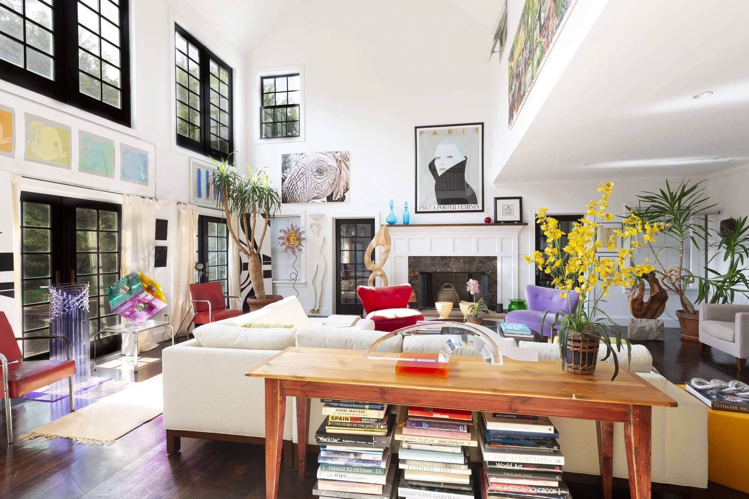 A brightly lit living room with large windows, colorful chairs, a white sofa, and various pieces of artwork and plants decorating the space.