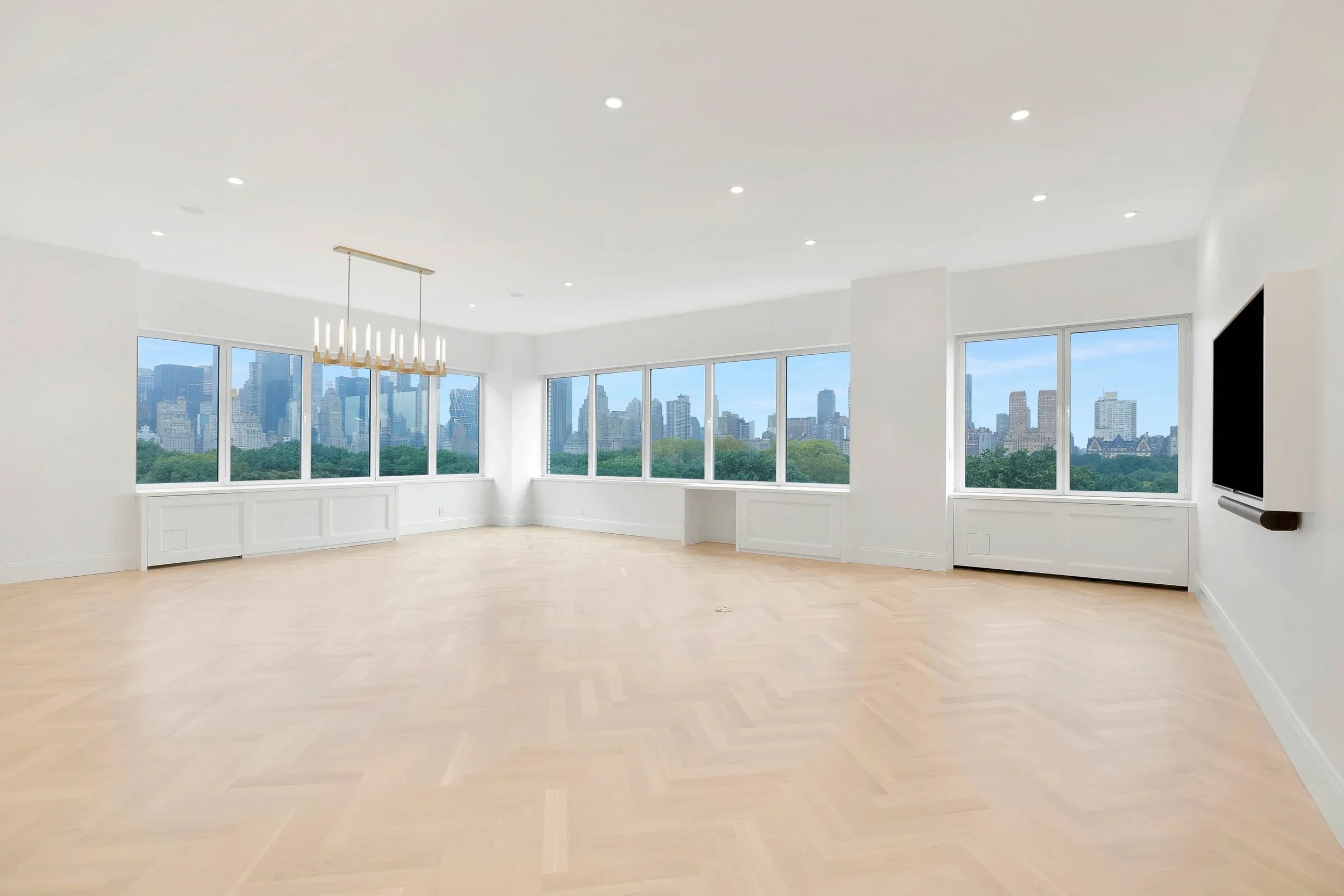 Empty bright room with large windows showing city skyline, white walls, light wood flooring, and a modern chandelier.