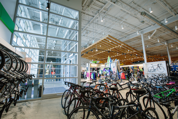 Windows at the store's entrance feature bright natural light, welcoming shoppers into the store. Cycling merchandise is on display in the front of the store, with LVL wood beem canopy in the distance.