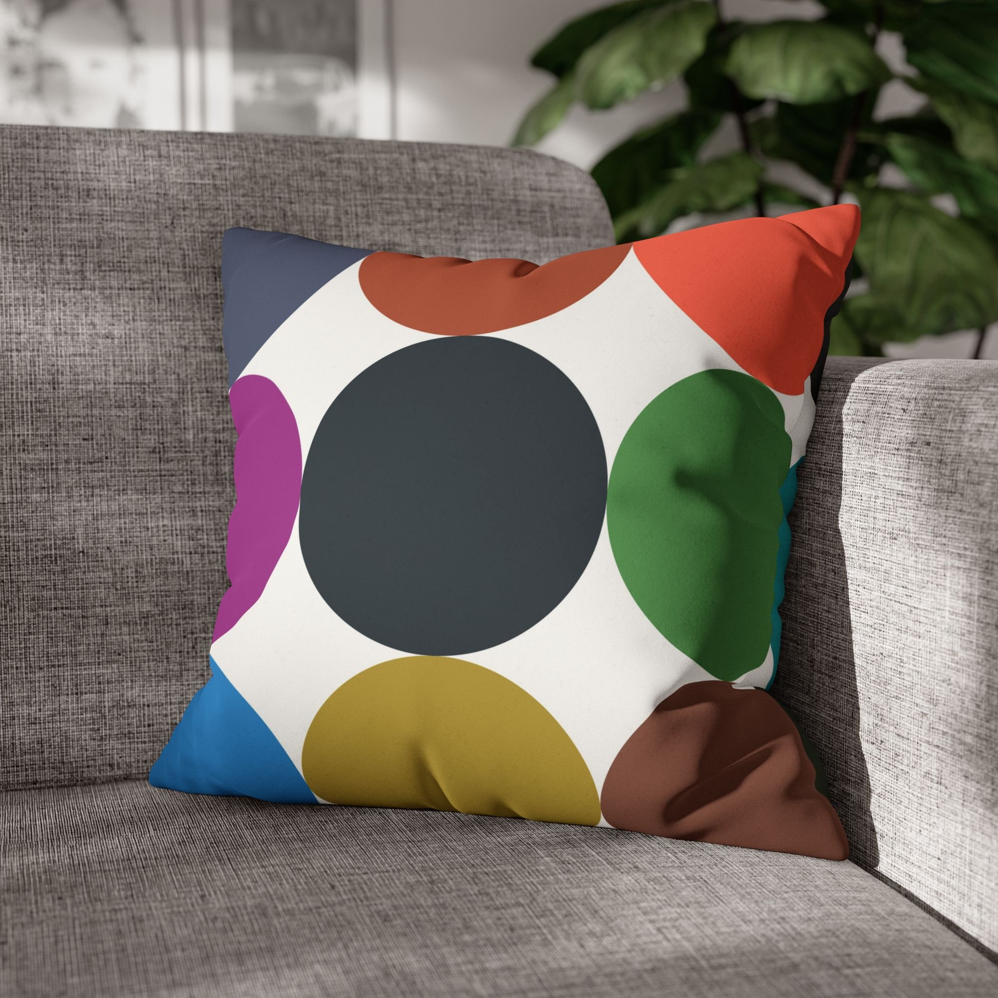 Colorful abstract-patterned throw pillow with large circles on a gray fabric sofa with a green plant in the background.