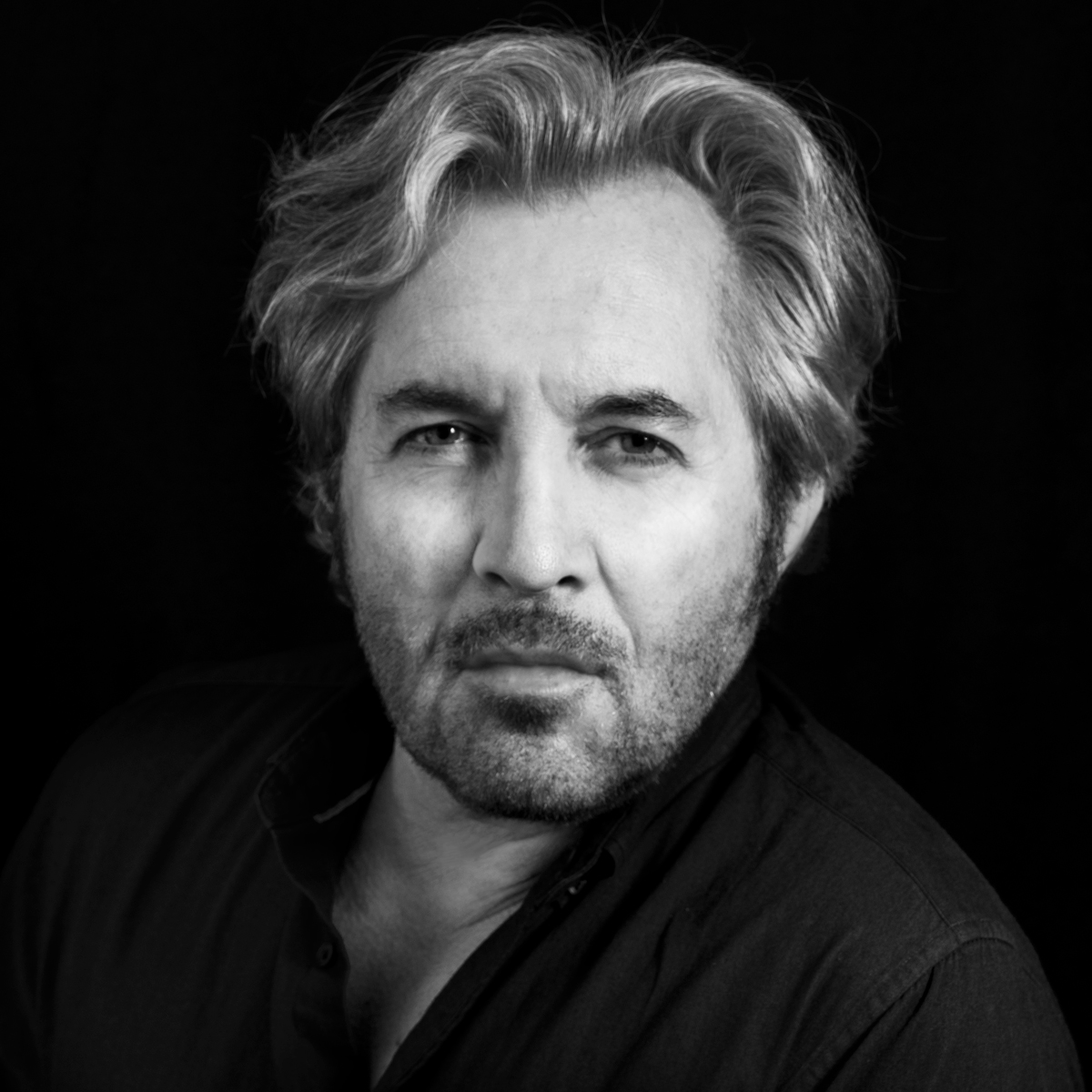 Black and white portrait of a man with gray hair and a beard, wearing a dark shirt, set against a dark background.