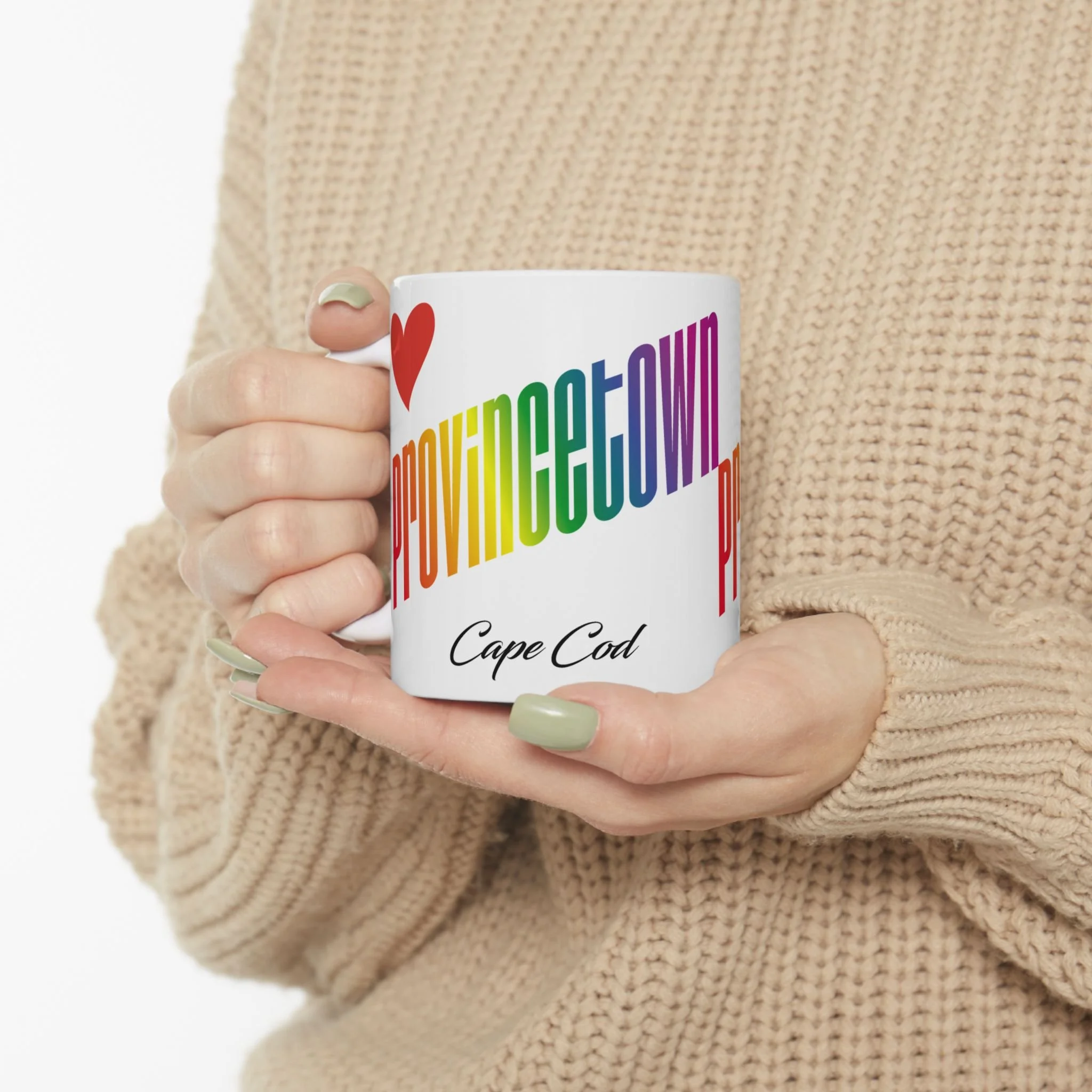 White mug with rainbow-colored lettering, held by a woman wearing a beige-colored sweater, tightly-cropped.