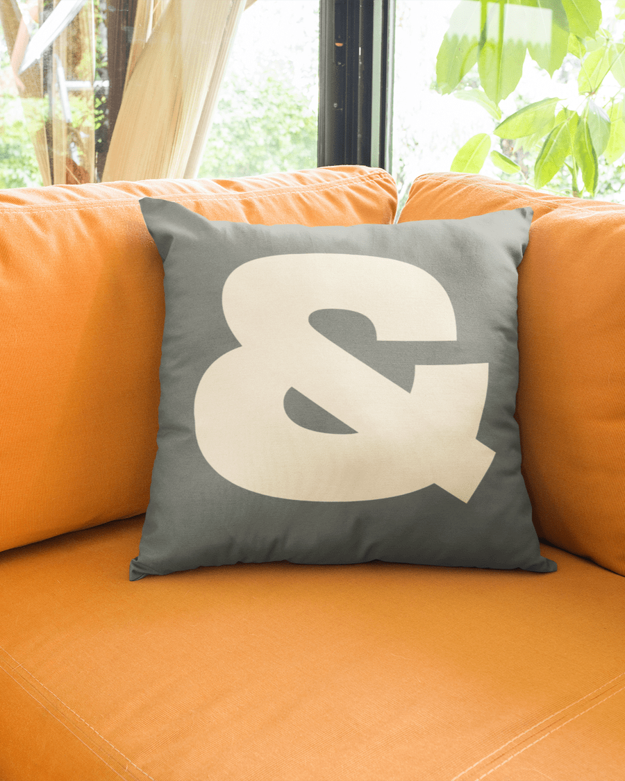 A square gray throw pillow with a large white ampersand symbol on it, placed on an orange couch near a window with green foliage outside.