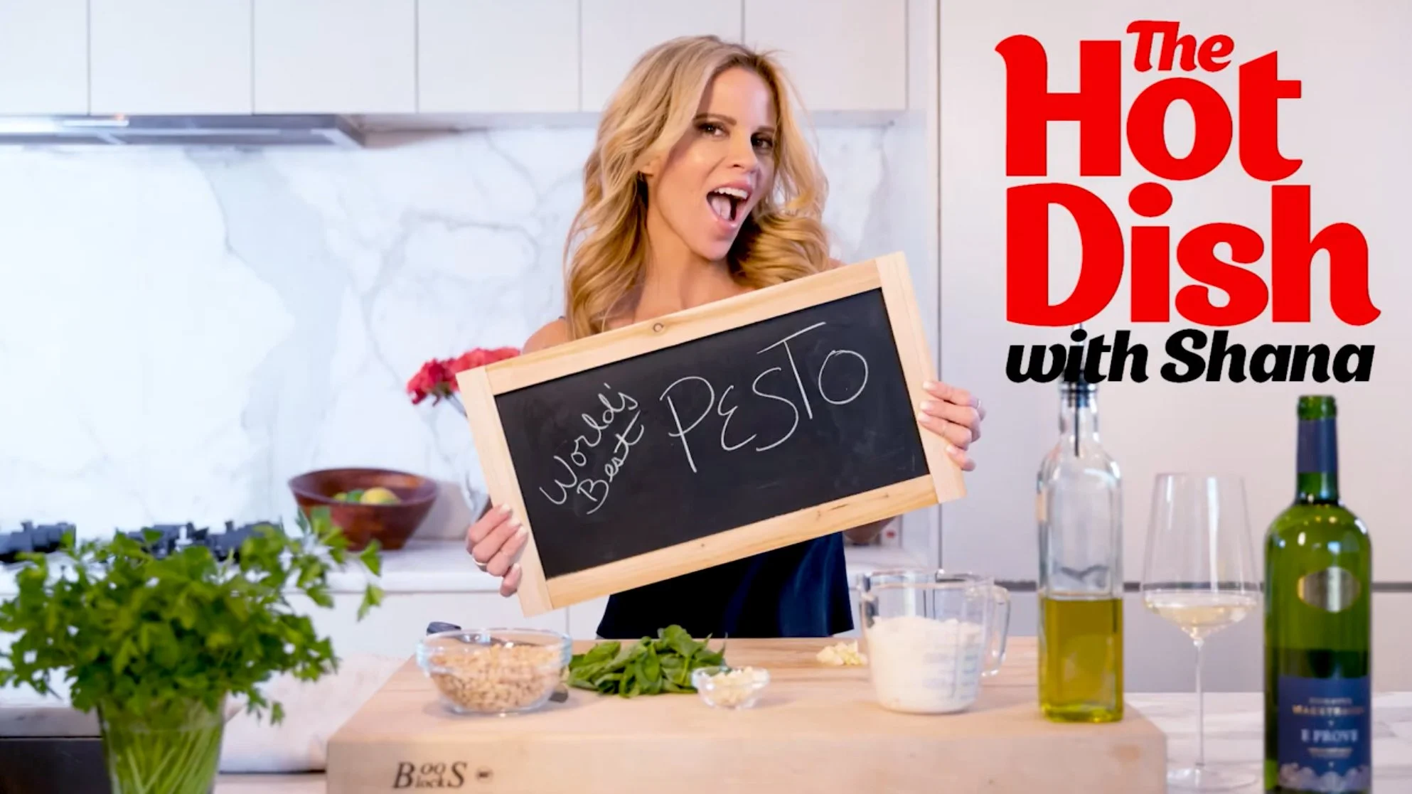 Video billboard, with a young blonde woman in a kitchen setting holding a small chalkboard with the word Pesto handwriten on it. The Hot Dish with Shana is in red nd black type in the upper right corner.