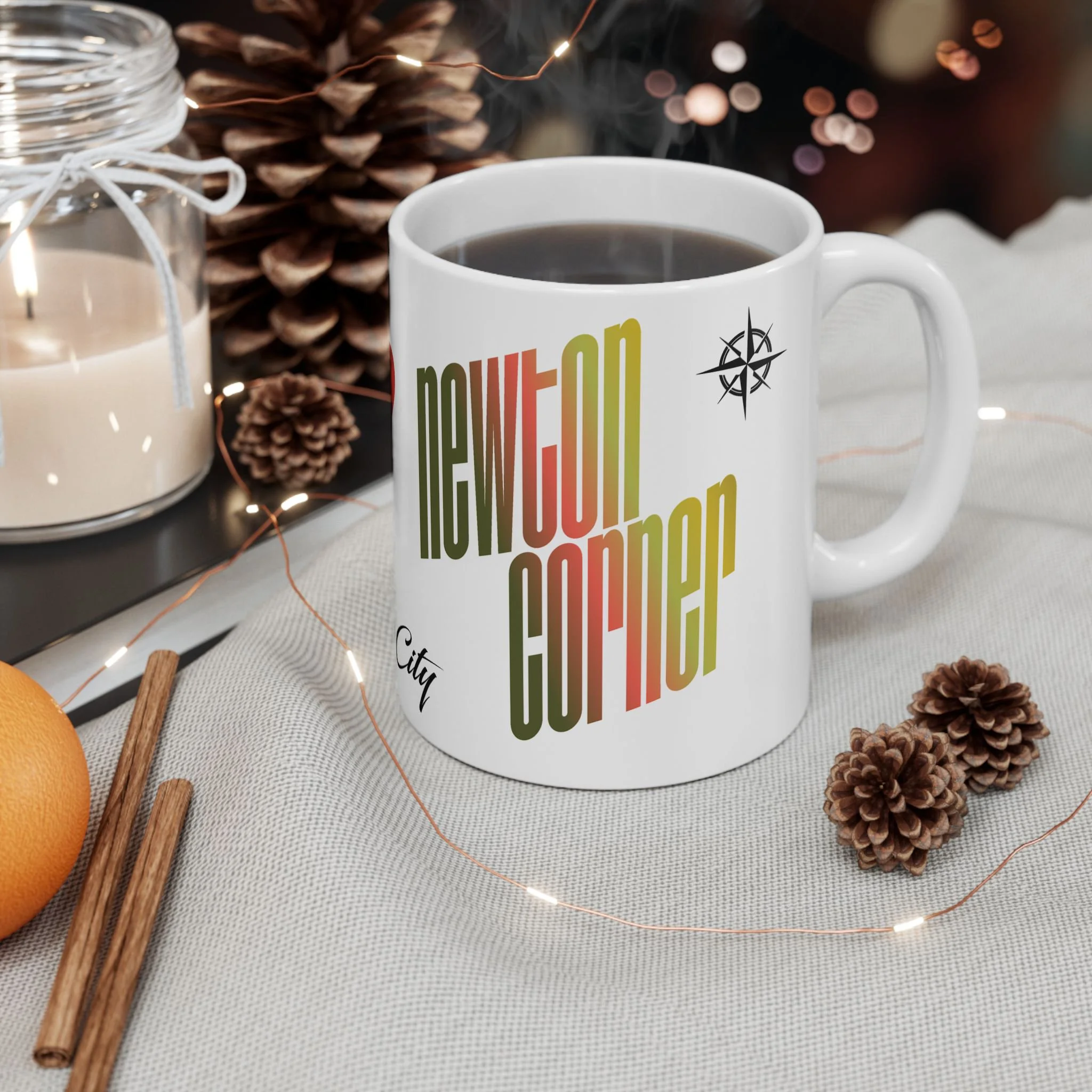 A white mug filled with coffee, with city and compass designs, placed on a table with pinecones, cinnamon sticks, an orange, a jar candle, and string lights around it, decorated for the holidays.
