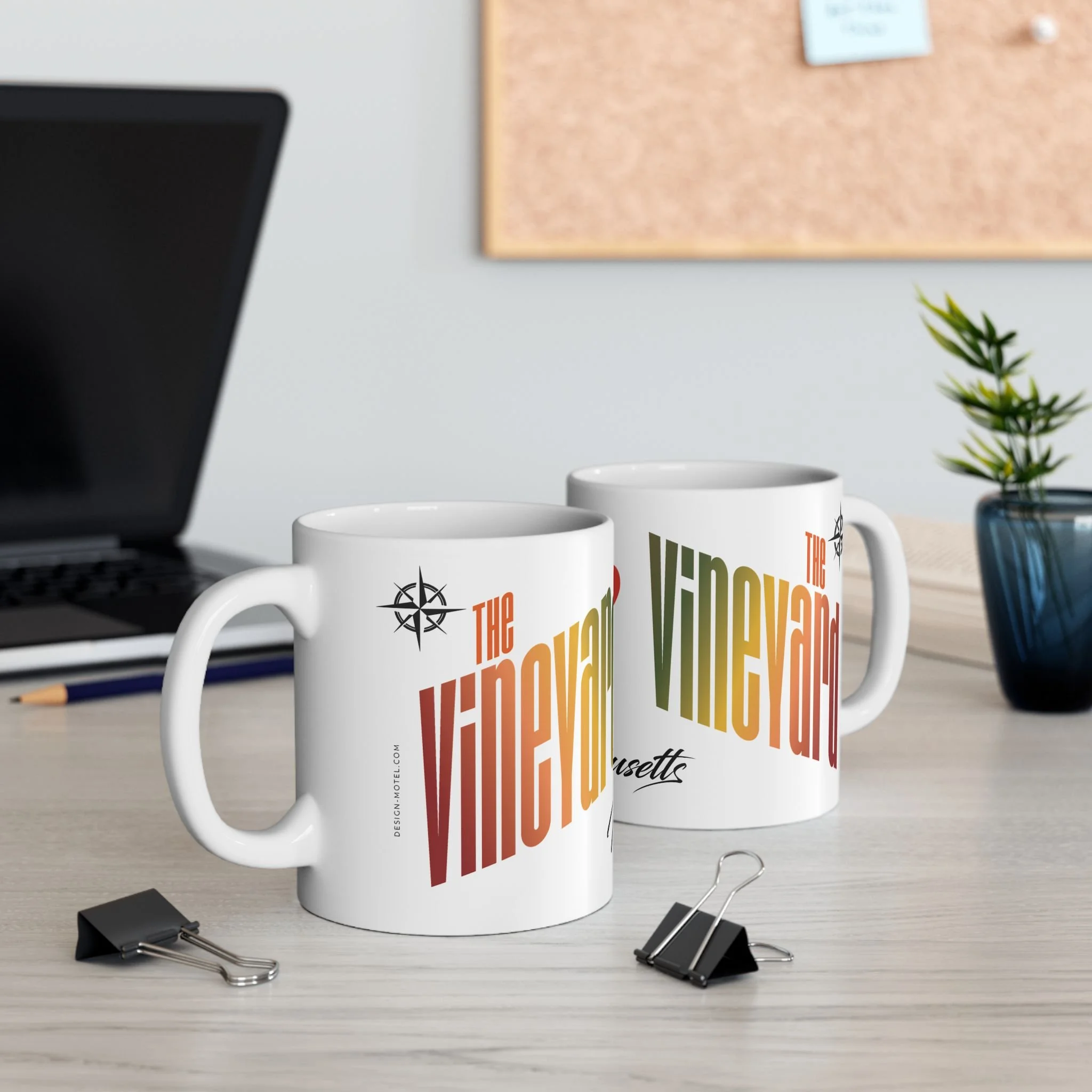 Two white coffee mugs with 'The Vineyard' logo and text on a desk, with black binder clips, a laptop, a pencil, a blue vase with a plant, and a corkboard in the background.