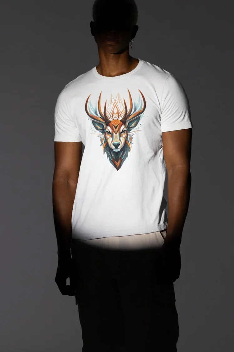 Animal spirit shirt featuring a stylized buck’s head on a white T-shirt, worn my a young Black man on a dark gray background. He is in shadow except for a spotlight on the shirt and his torso.
