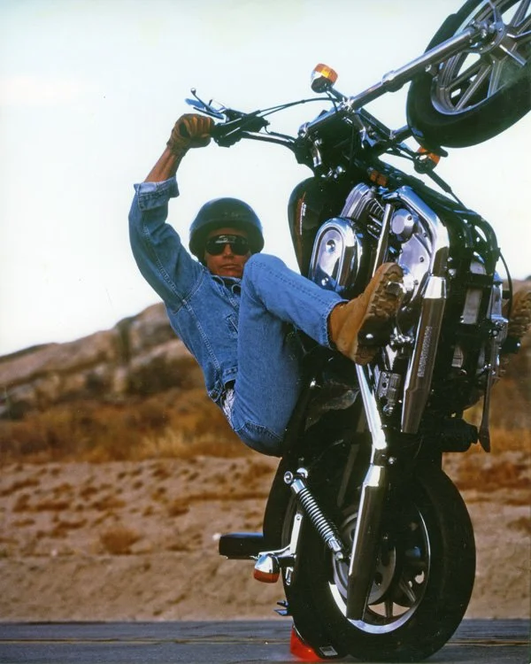 Photo of Doug Domokos riding a motorcycle on only the back wheel while looking at the camera. The motorcycle is near-vertical with scrubby California landscape in the background.
