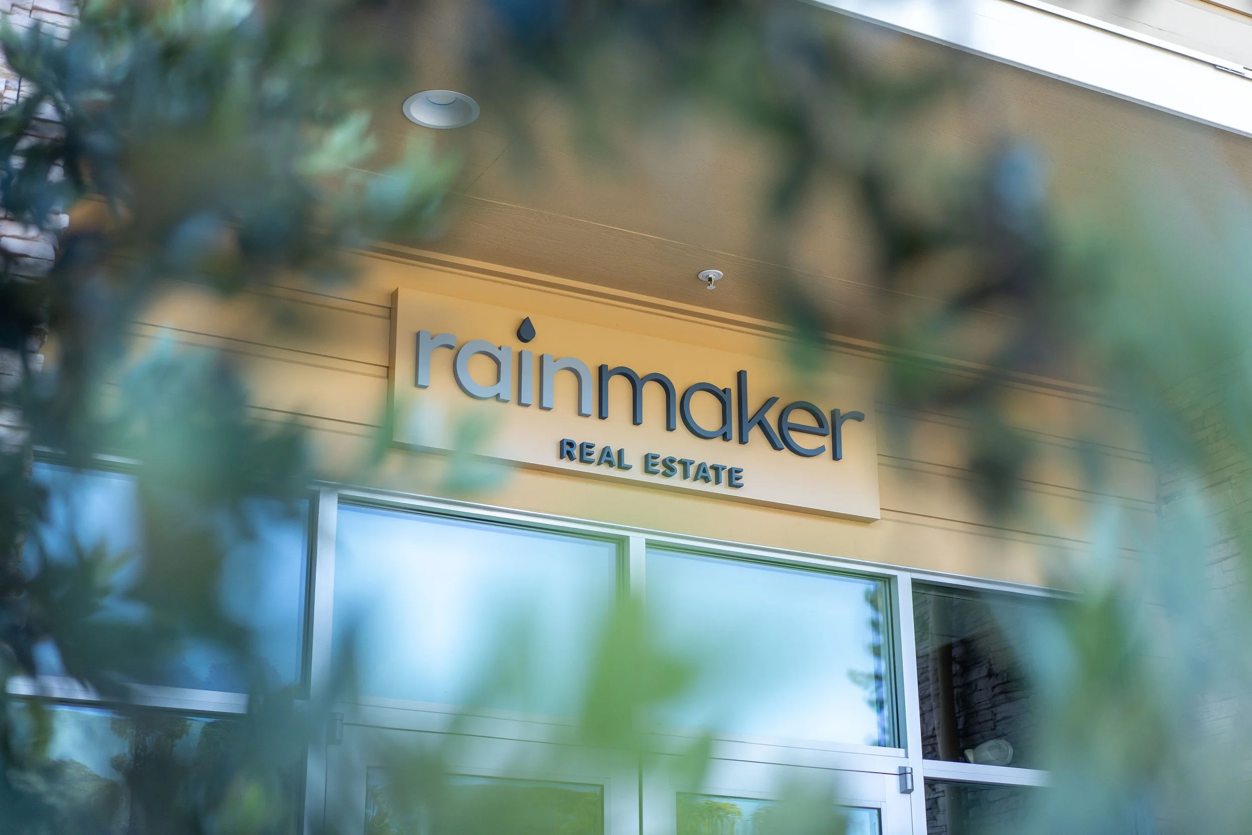 Rainmaker Real Estate office sign on a beige building, partially obscured by green foliage.