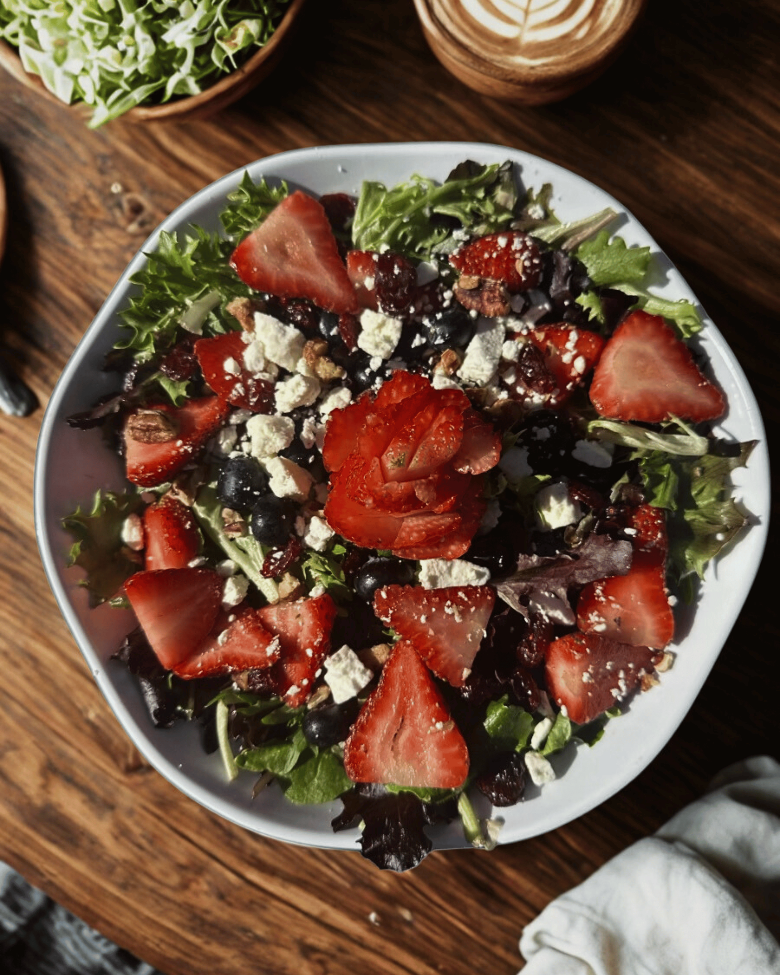 A fresh mixed green salad with sliced strawberries, blueberries, crumbled cheese, and chopped nuts in a white bowl on a wooden table.