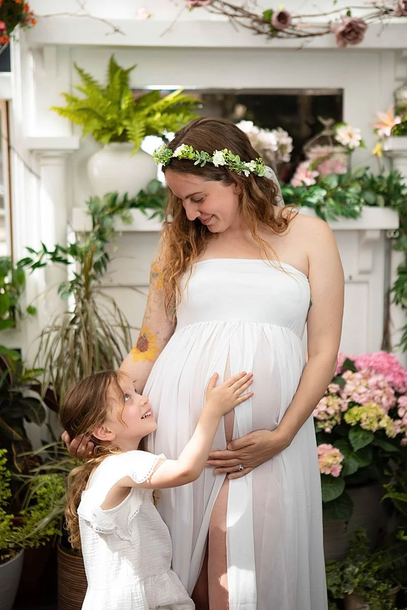 Expecting mother in a white strapless maternity dress in a floral crown posing for portraits with her young daughter. The little girl has her hand on her mom’s belly looking up at her. Maternity photographer in Chesterfield Virginia