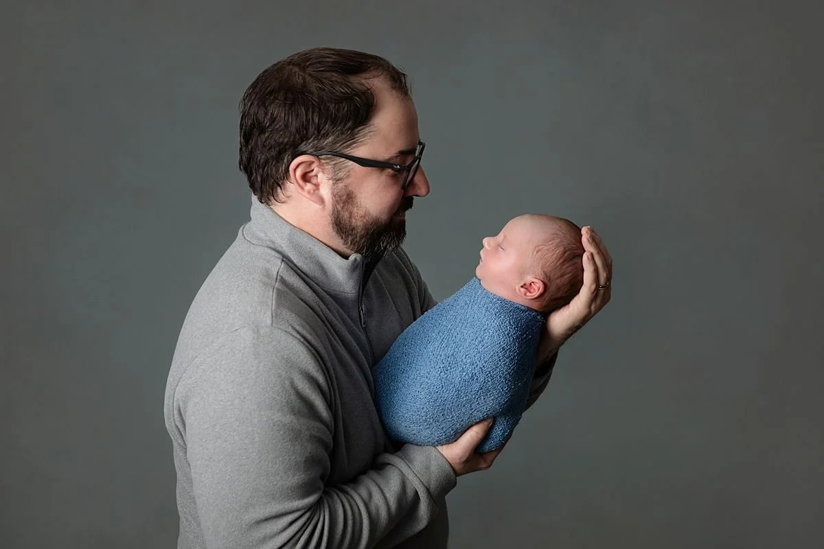 Chesterfield Virginia Newborn Photographer, Dad holding his newborn baby during a photo shoot in Chesterfield VA baby is wrapped in blue with a gray background