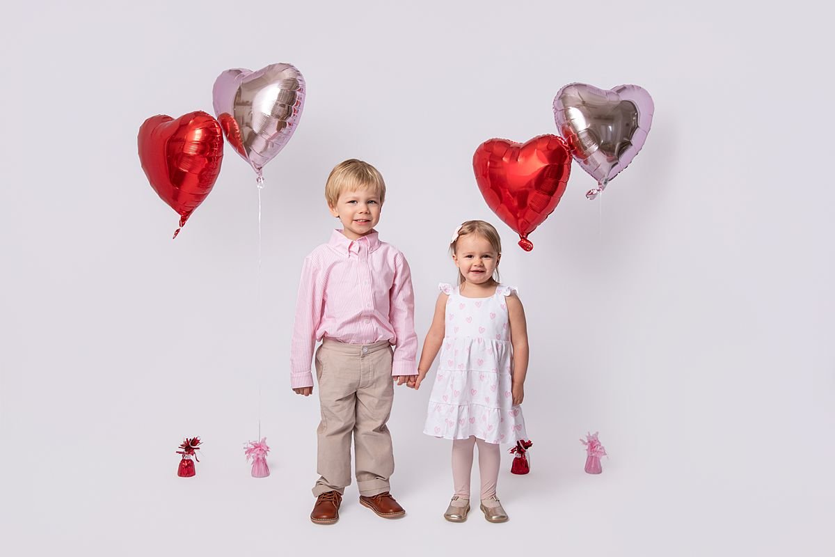 Young brother and sister holding hands smiling with heart balloons behind them photographed by Sarah Kane Photography near Richmond VA