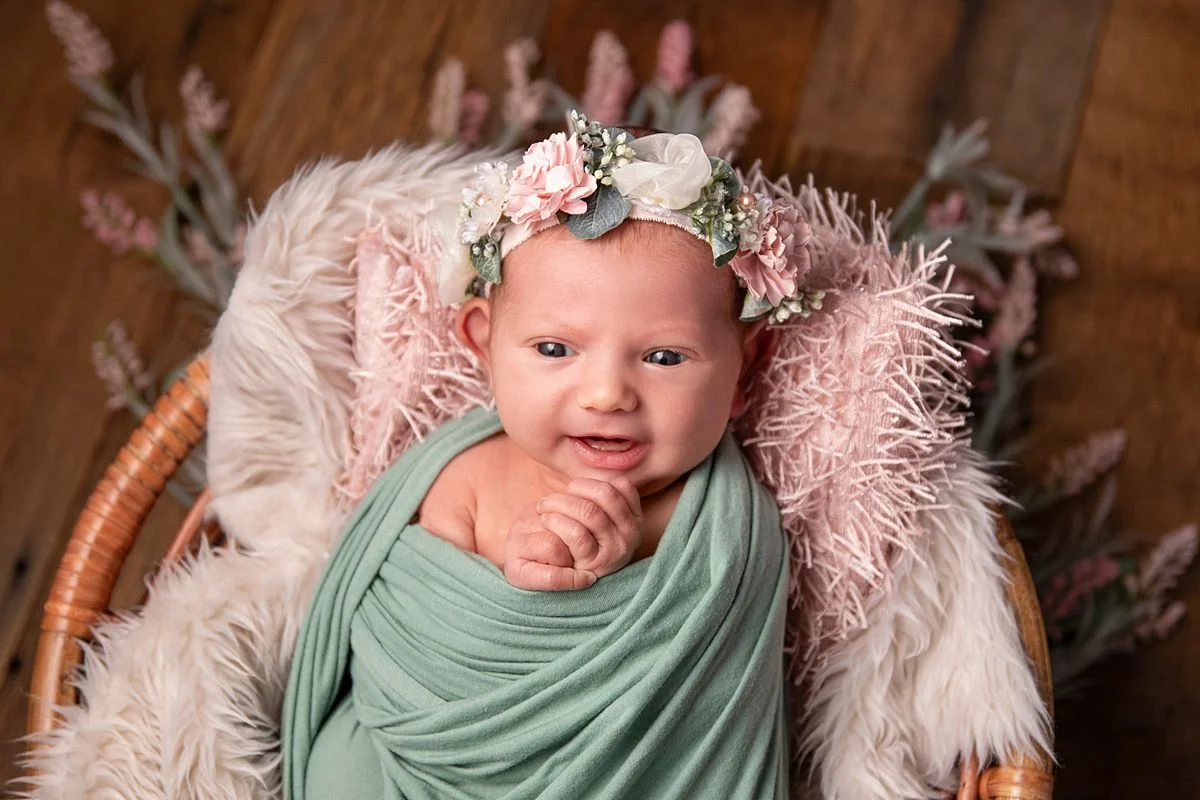 Alert newborn girl in sage green swaddle and pink floral headband smiling in wicker basket, Sarah Kane Photography