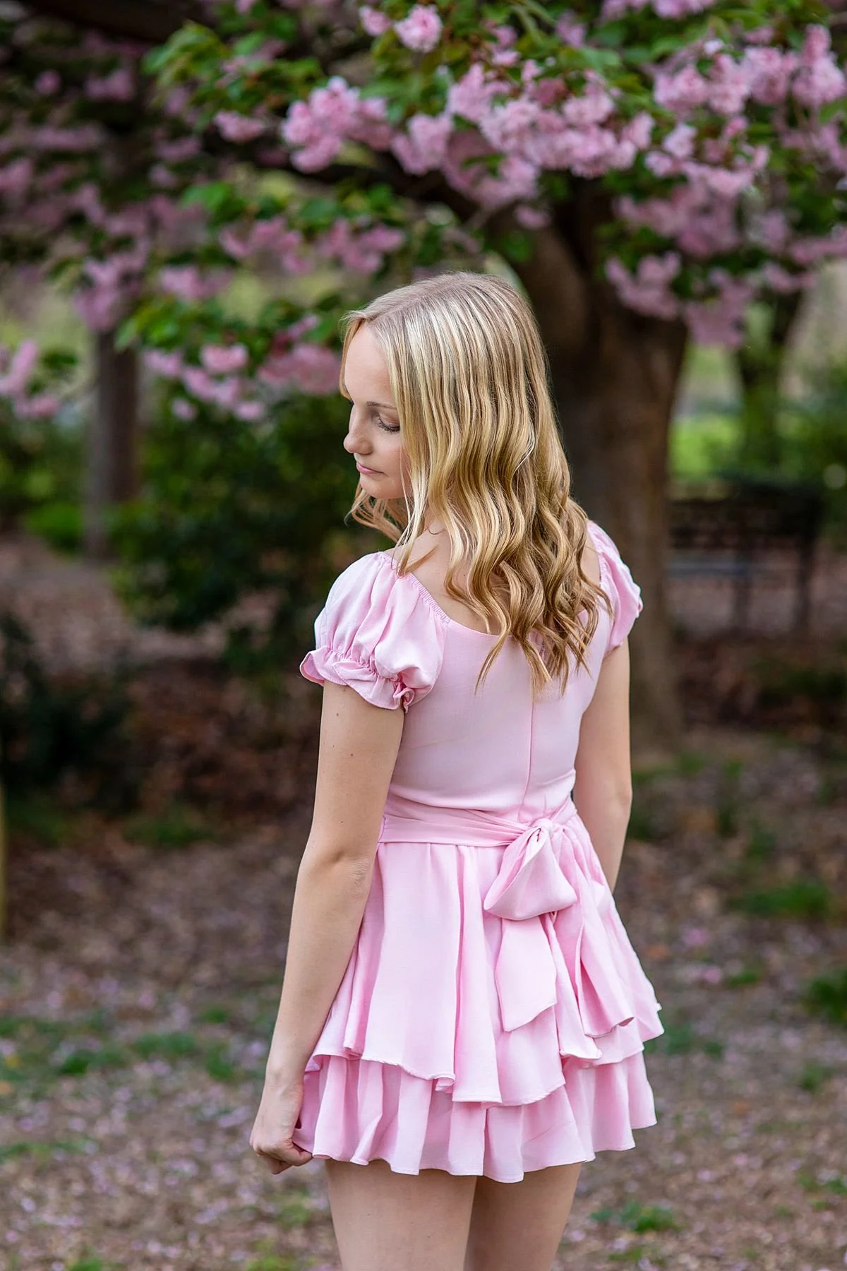 Back turned artistic senior portrait of a girl in a pink ruffle dress standing in front of lush blooming pink trees in spring, near Richmond, Virginia, Sarah Kane Photography, Chesterfield VA