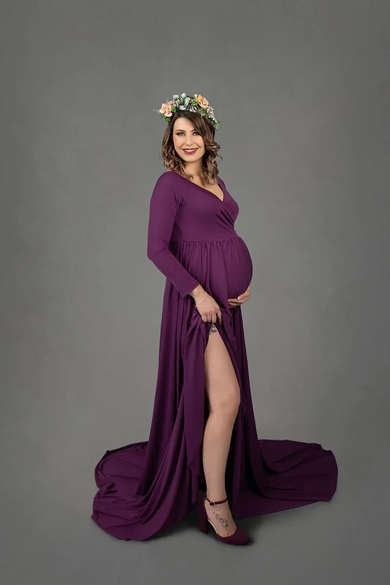 Pregnant woman in a purple long maternity gown wearing a flower crown with a grey background for maternity pictures near Richmond Virginia