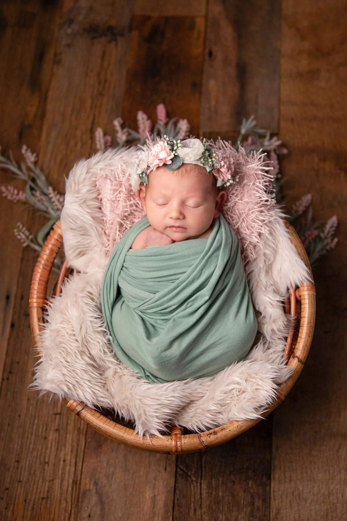 Sleeping newborn in sage green wrap and pink floral headband in round wicker basket, Sarah Kane Photography