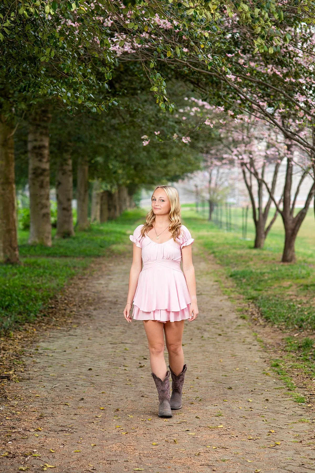Blonde high school senior in a pink dress and cowboy boots walks toward the camera down a stunning tree lined path during a spring senior portrait session near Richmond, Virginia, photographed by Sarah Kane Photography