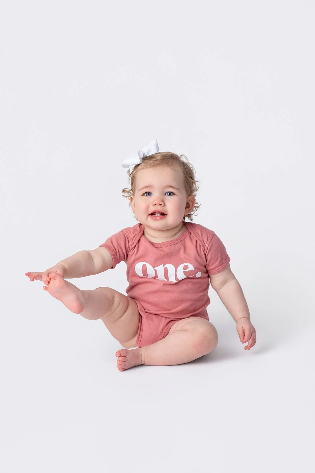 One year old baby holding her foot in the air with a white bow, Sarah Kane Photography