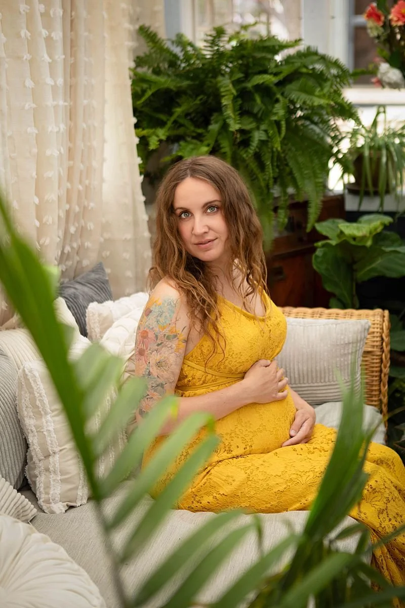 maternity portraits of an expecting mother in a golden yellow dress sitting on a day bed in a cottage filed with plants and natural light.  Located in Chesterfield Virginia close to Richmond.