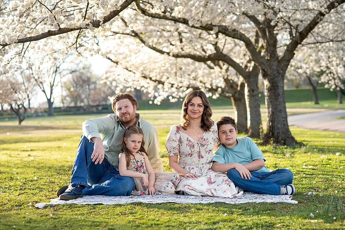 a family of four sitting together on a white lace blanket during a spring family portrait session in Richmond Virginia. Dad with a red beard in a sage green shirt and jeans, mom in a floral dress, a young daughter  and son under a cherry blossom tree