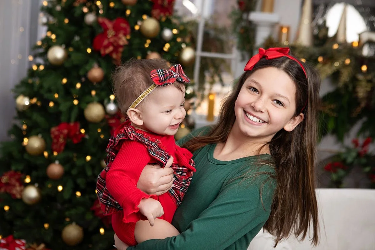 A holiday picture of a older girl dressed in green holding her baby sister dressed in red plaid in front of a Christmas tree in Chesterfield Virginia