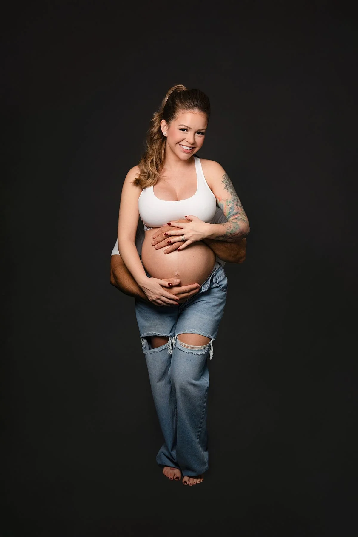 Pregnant woman in white top and jeans smiling holding her belly with her partners arms around her, Sarah Kane Photography