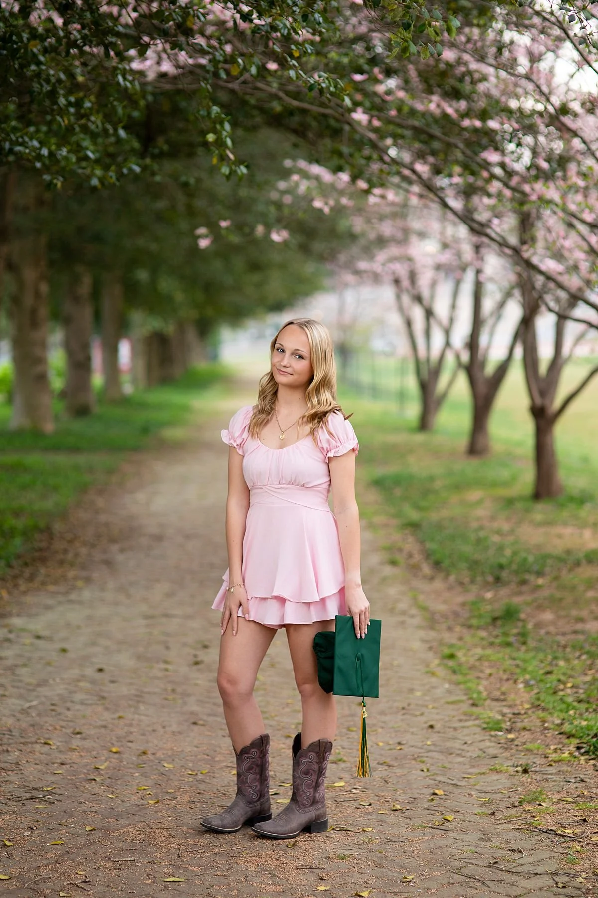 Senior girl in a pink ruffled dress and brown cowboy boots walks along a canopied tree lined path during a senior portrait session holding a graduation cap, near Richmond, Virginia, Sarah Kane Photography, Chesterfield, VA